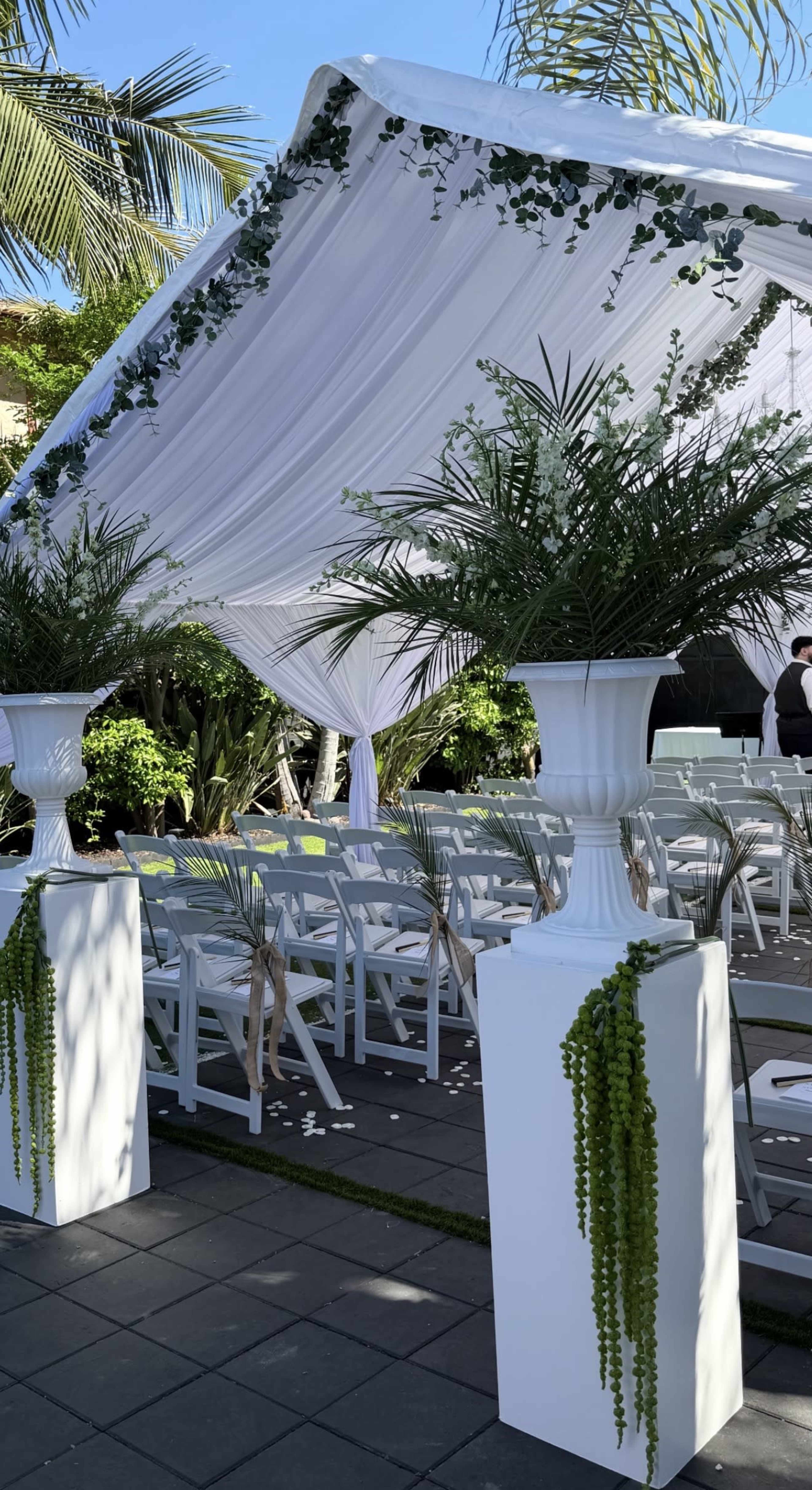 The image shows a wedding venue featuring a white canopy adorned with greenery, white chairs arranged for guests, and decorative planters on either side.