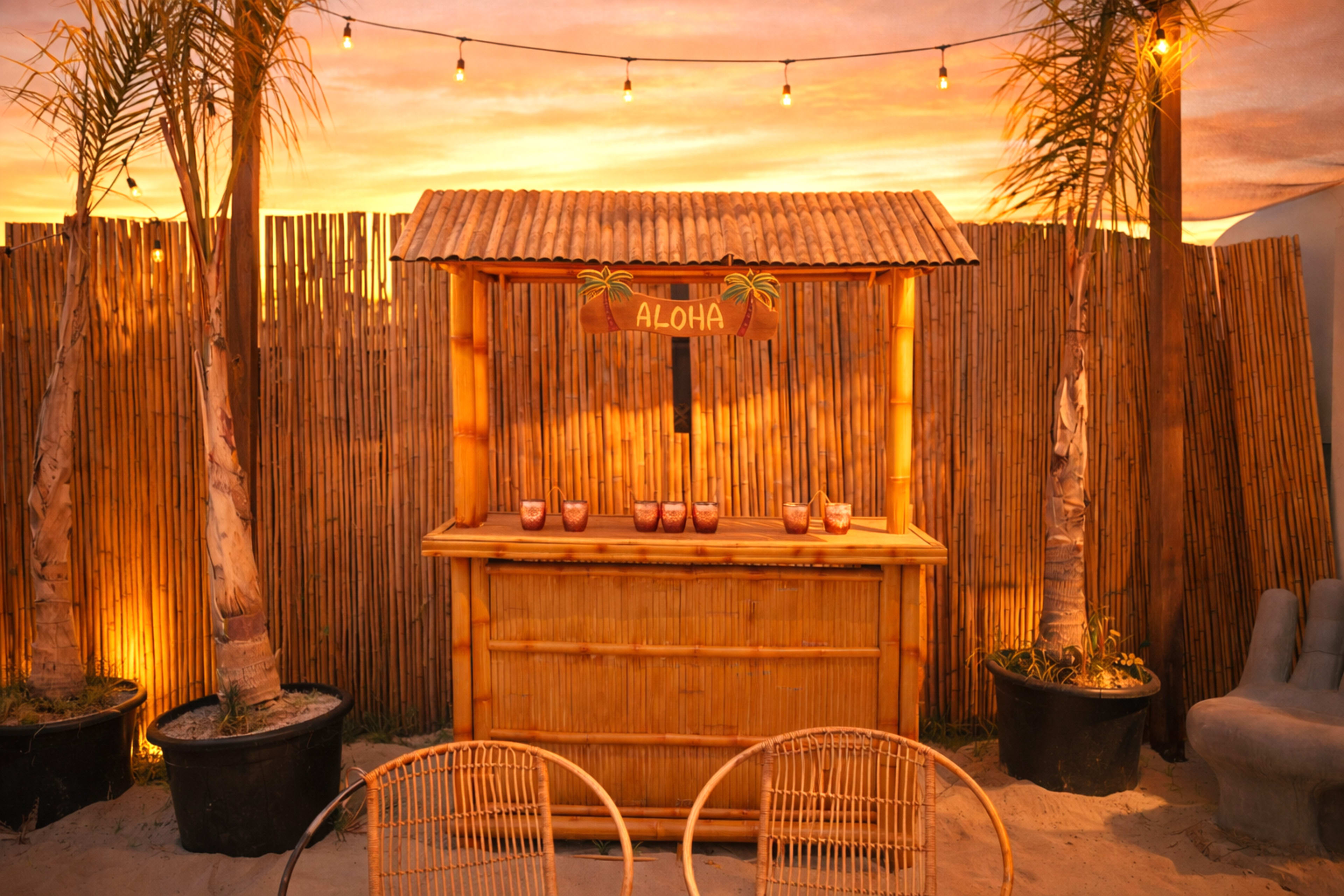 A bamboo bar with the word "ALOHA" is set against a wooden fence, surrounded by potted palms, under string lights with a sunset in the background.