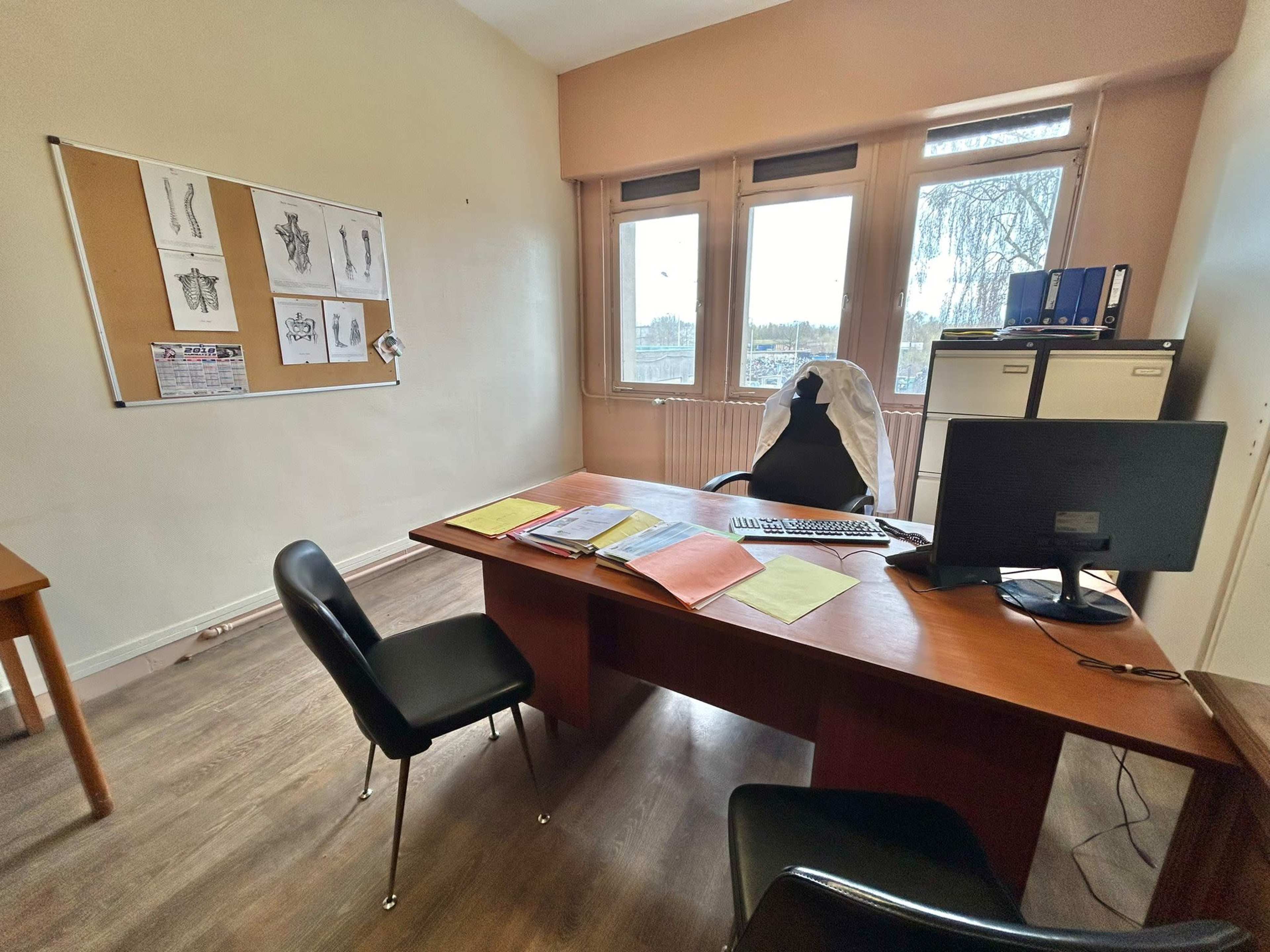 A spacious office features a large wooden desk with a computer and various documents, alongside a chair, two black chairs, and windows that provide natural light.