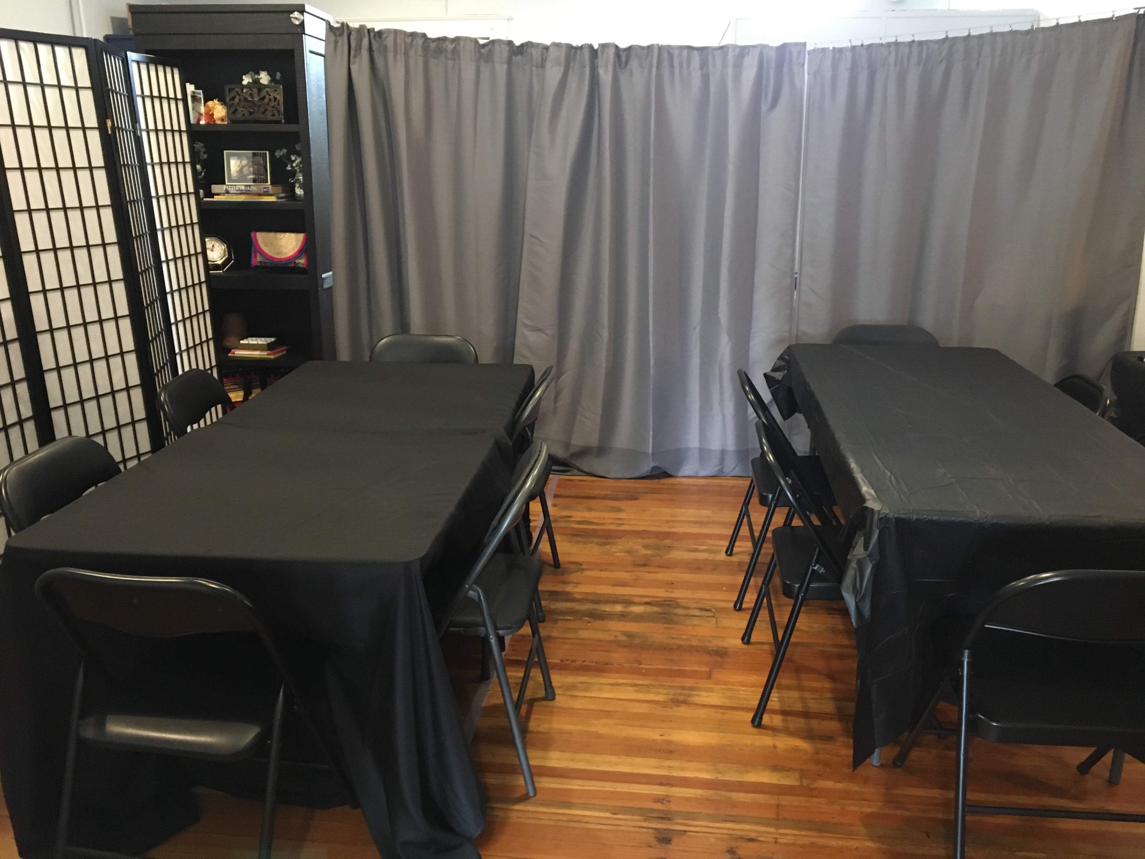 A room with two sets of black-draped tables separated by a gray curtain, accompanied by black chairs and wooden flooring.