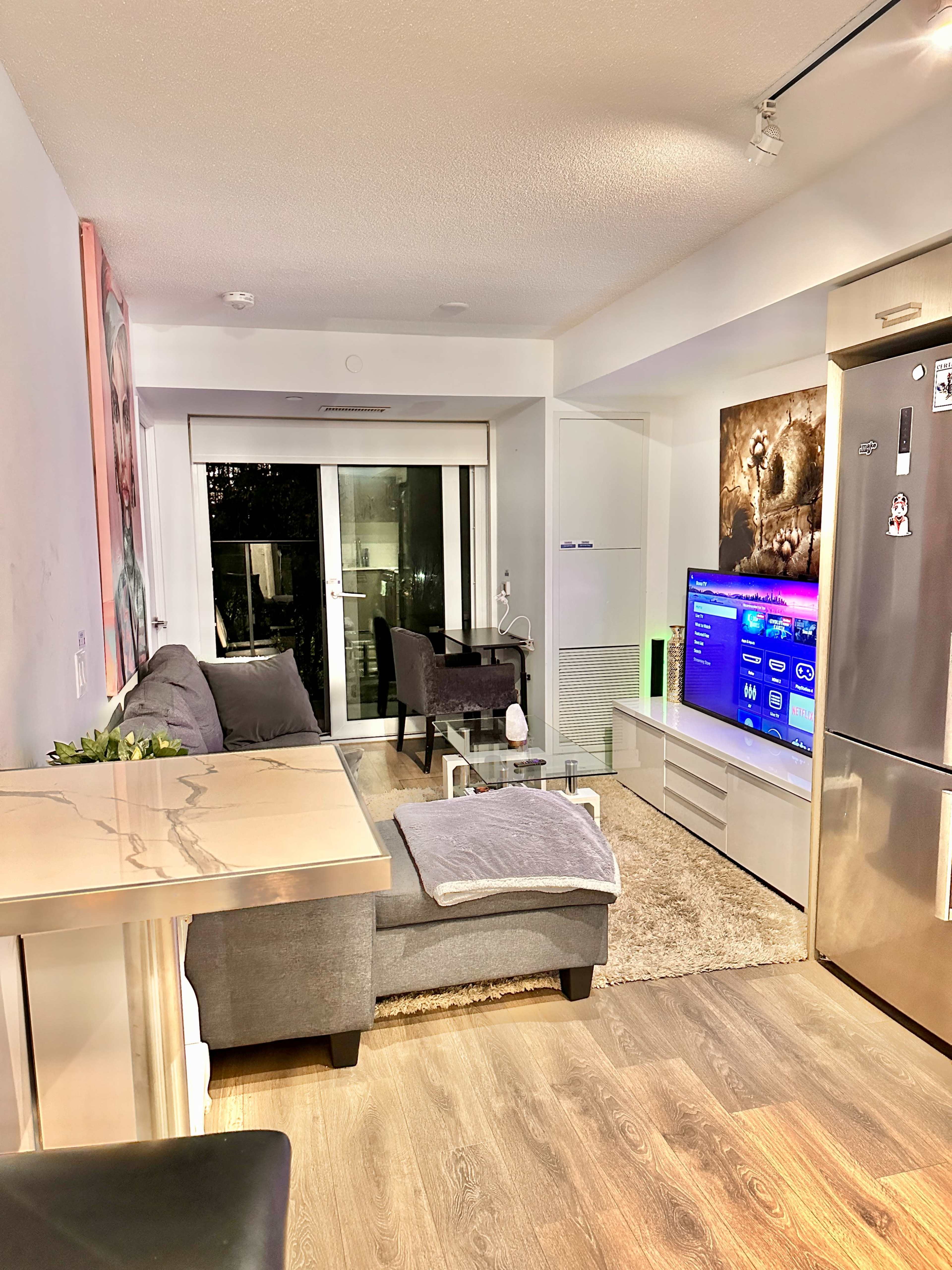 The image depicts a modern living room with a gray sofa, a flat-screen TV on a white media unit, and large windows leading to a balcony.