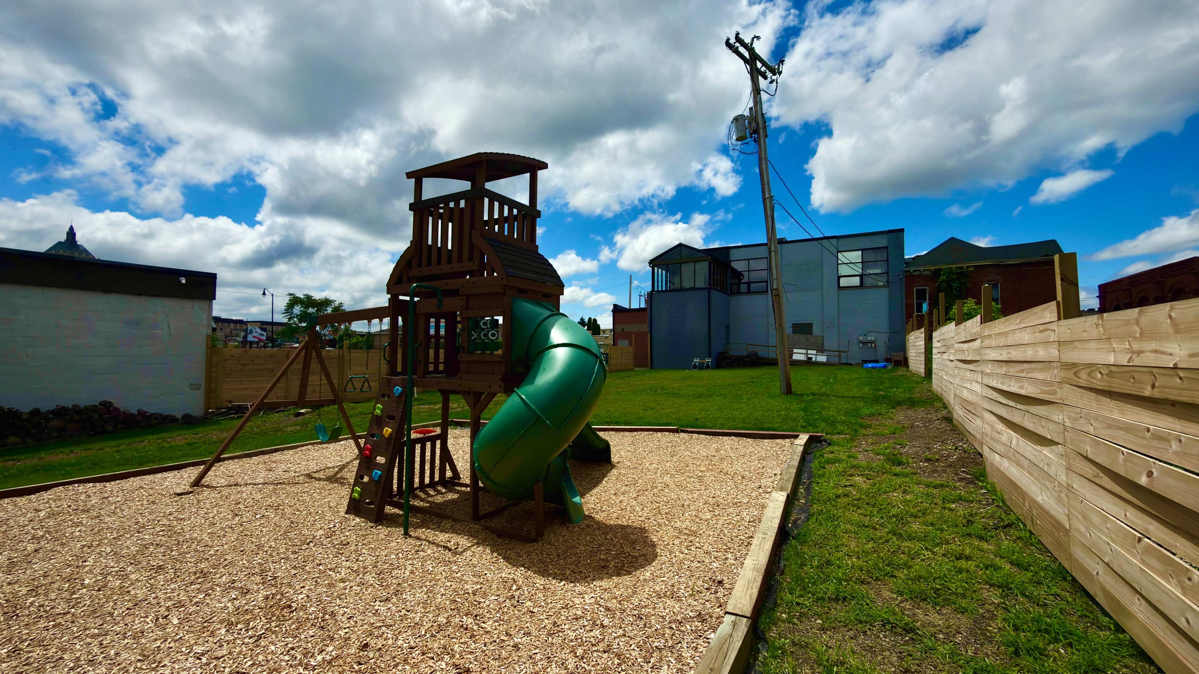 A wooden playground structure with a green slide is situated on a gravel surface in a park-like area, with a backdrop of buildings and power lines under a partly cloudy sky.