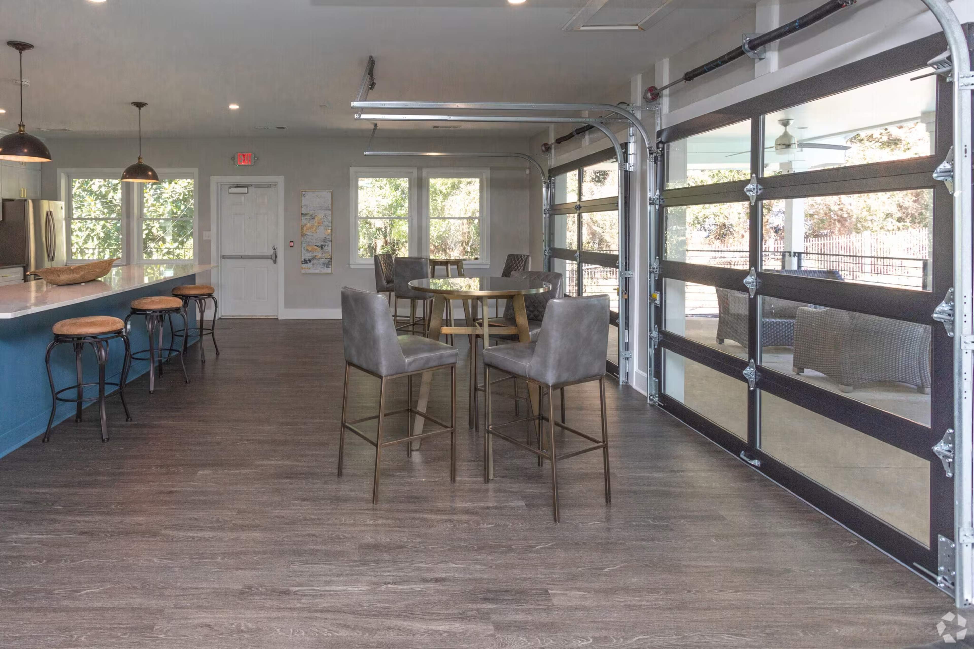 The image shows a modern indoor space with a bar counter, high chairs, tables, and large glass garage doors leading to an outdoor area.