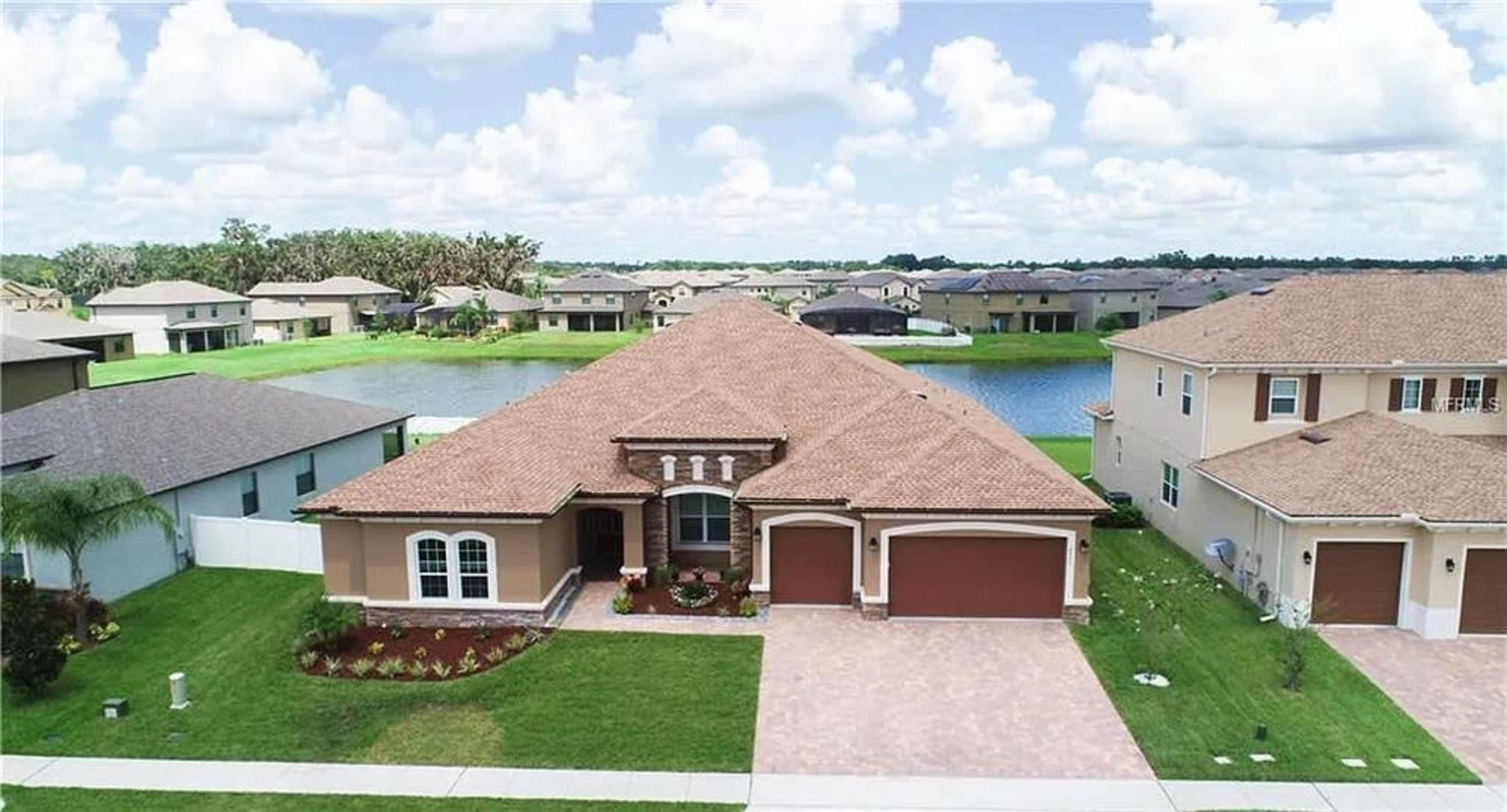 The image shows a single-story house with a brown tiled roof, front garden, and two garage doors, situated near a calm body of water and surrounded by other residential buildings.