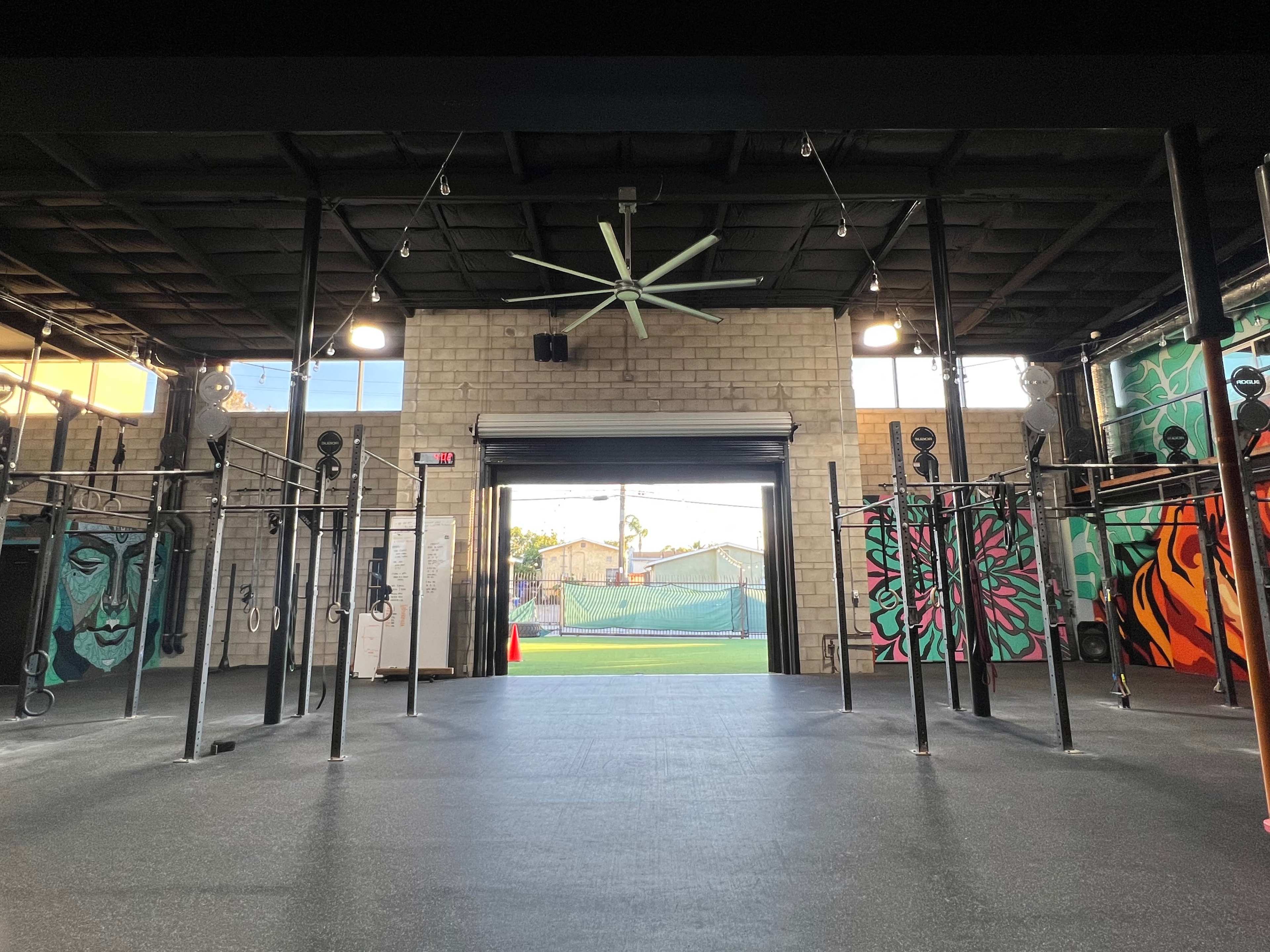 The image shows a gym interior with mounted exercise equipment and colorful murals on the walls, leading to an open area with a tennis court visible in the background.