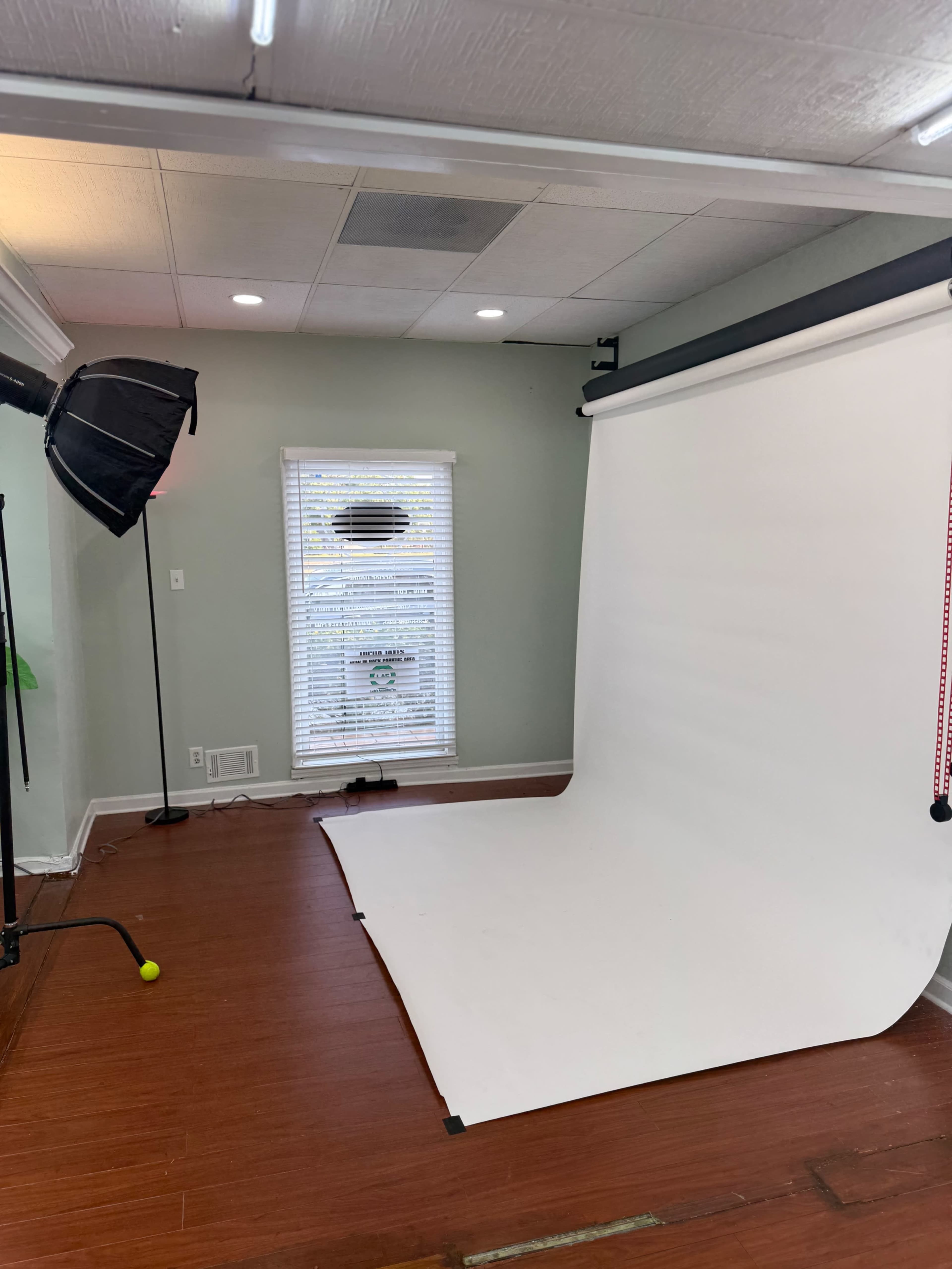 The image shows a studio space featuring a white backdrop setup, a softbox light, and a window with blinds.