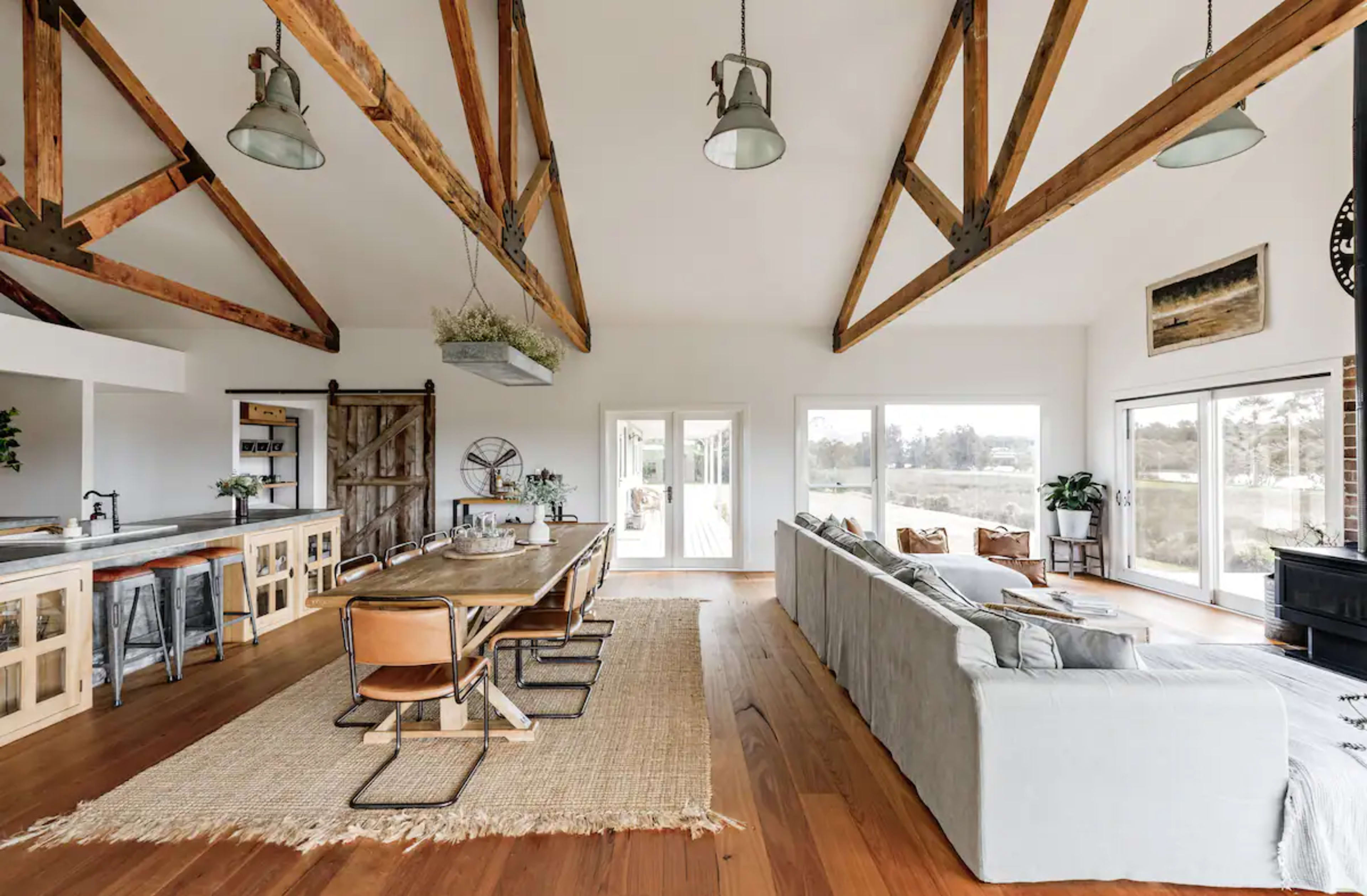 A spacious living area featuring exposed wooden beams, a large dining table, and large windows overlooking a natural landscape.