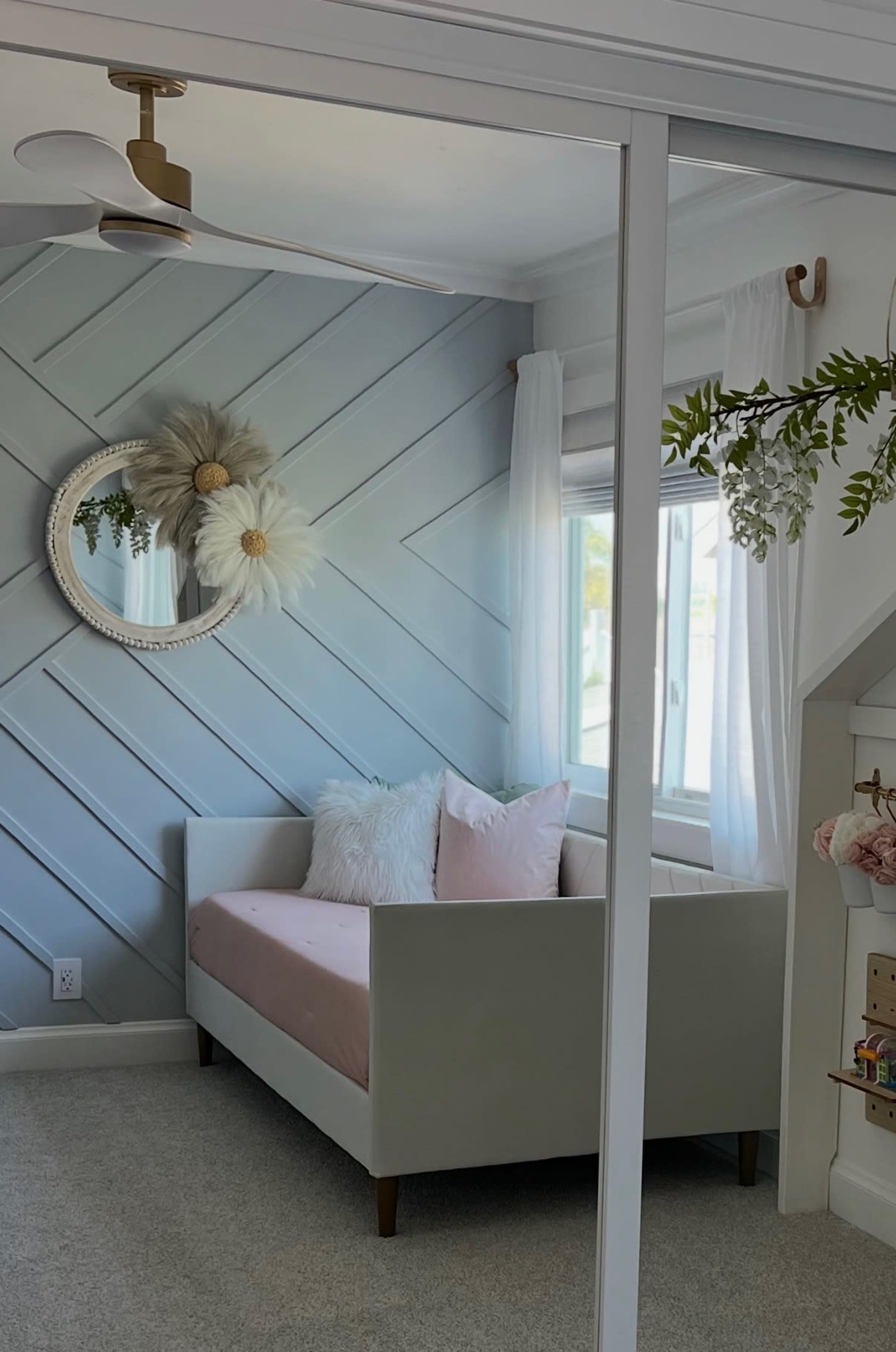 The image shows a light gray daybed with pink cushions positioned against a diagonal striped wall and a window adorned with white curtains, reflecting a serene interior design.