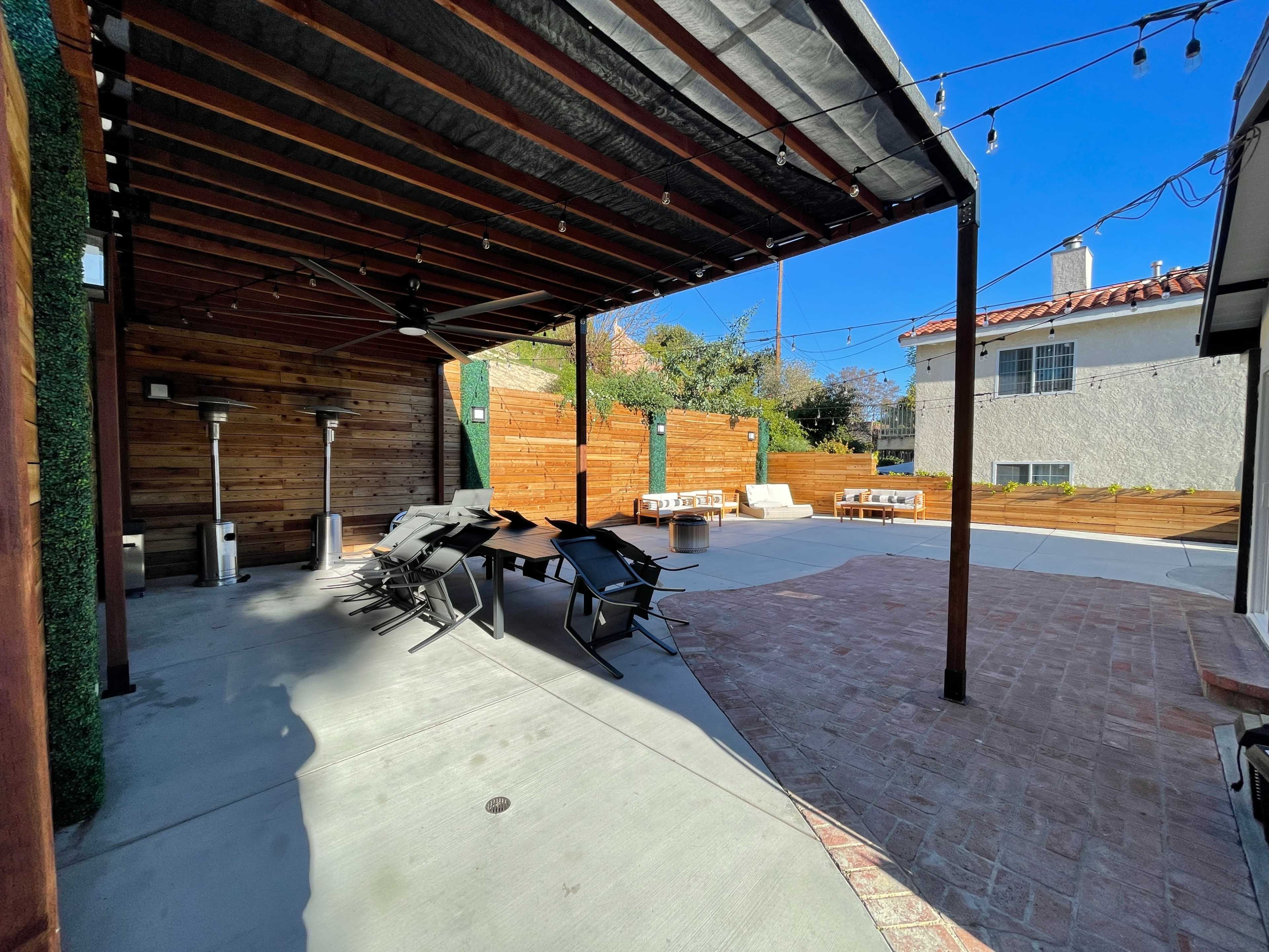 The image shows a spacious outdoor patio area with seating arrangements, metal heaters, and wooden fences under a clear blue sky.