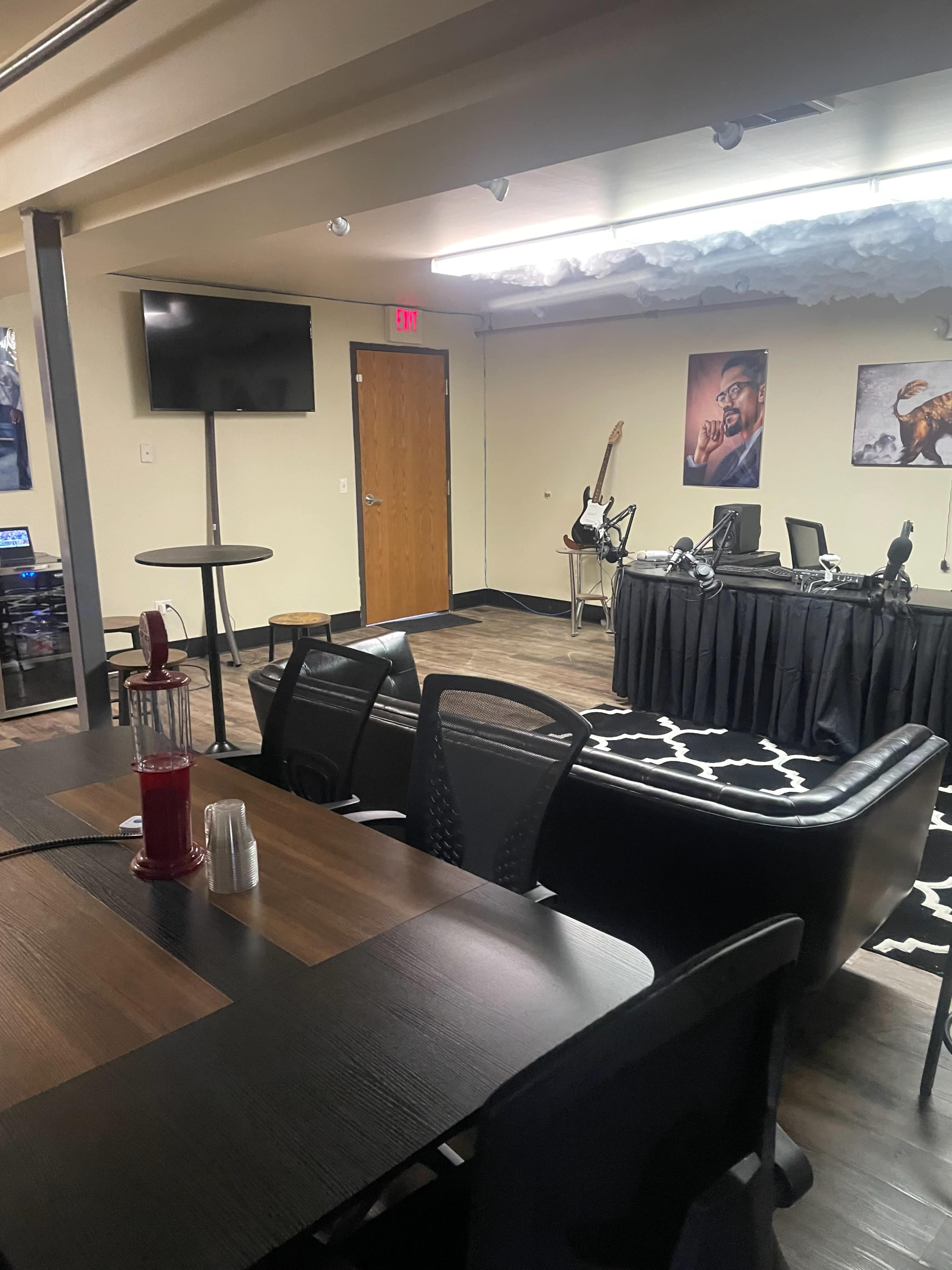 An indoor space featuring a seating area with black chairs and a table, a television on the wall, and a corner stage with musical instruments and microphones.