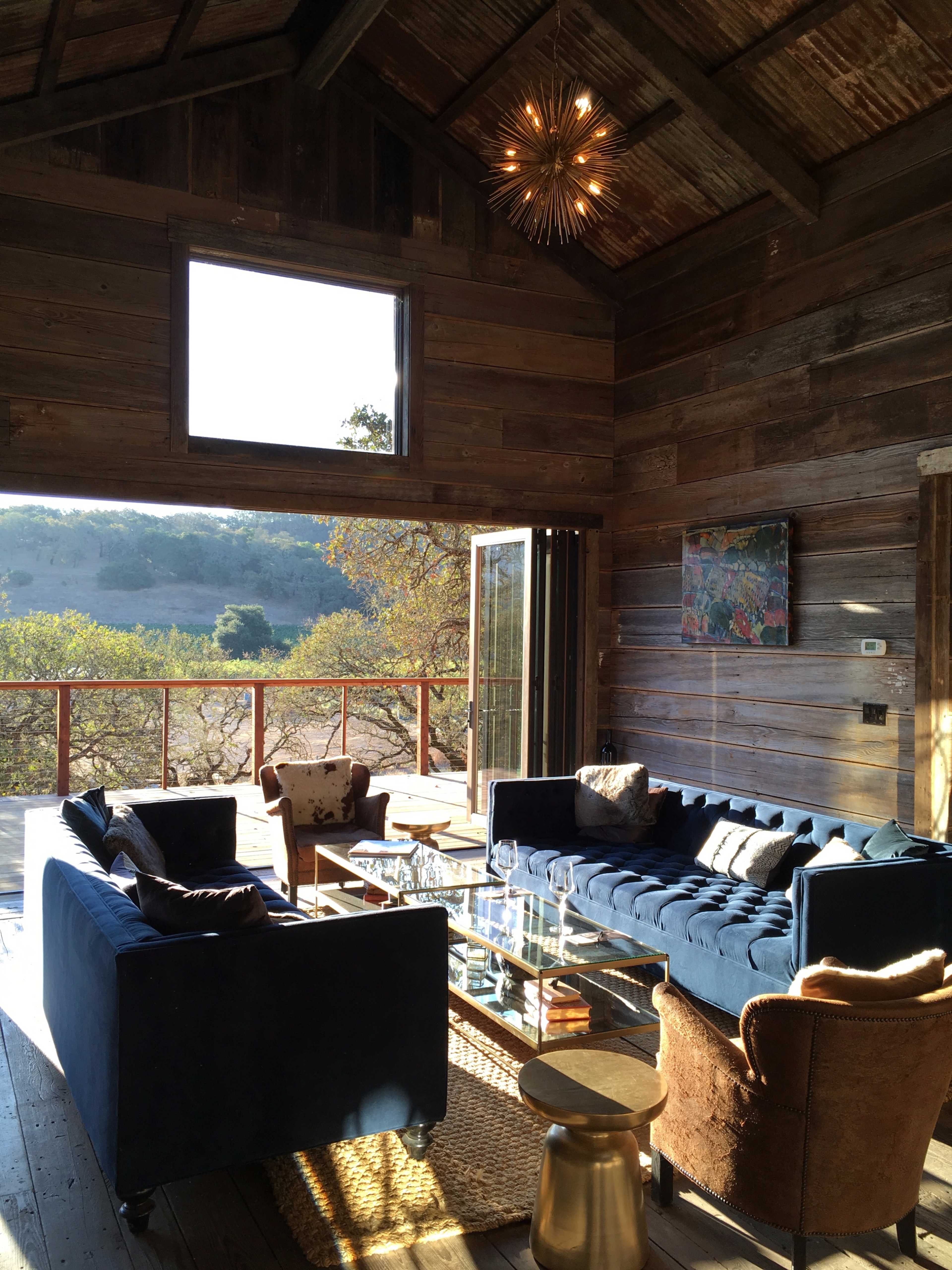 A modern, rustic living room features blue sofas, a glass coffee table, and large windows providing a view of the outdoors.