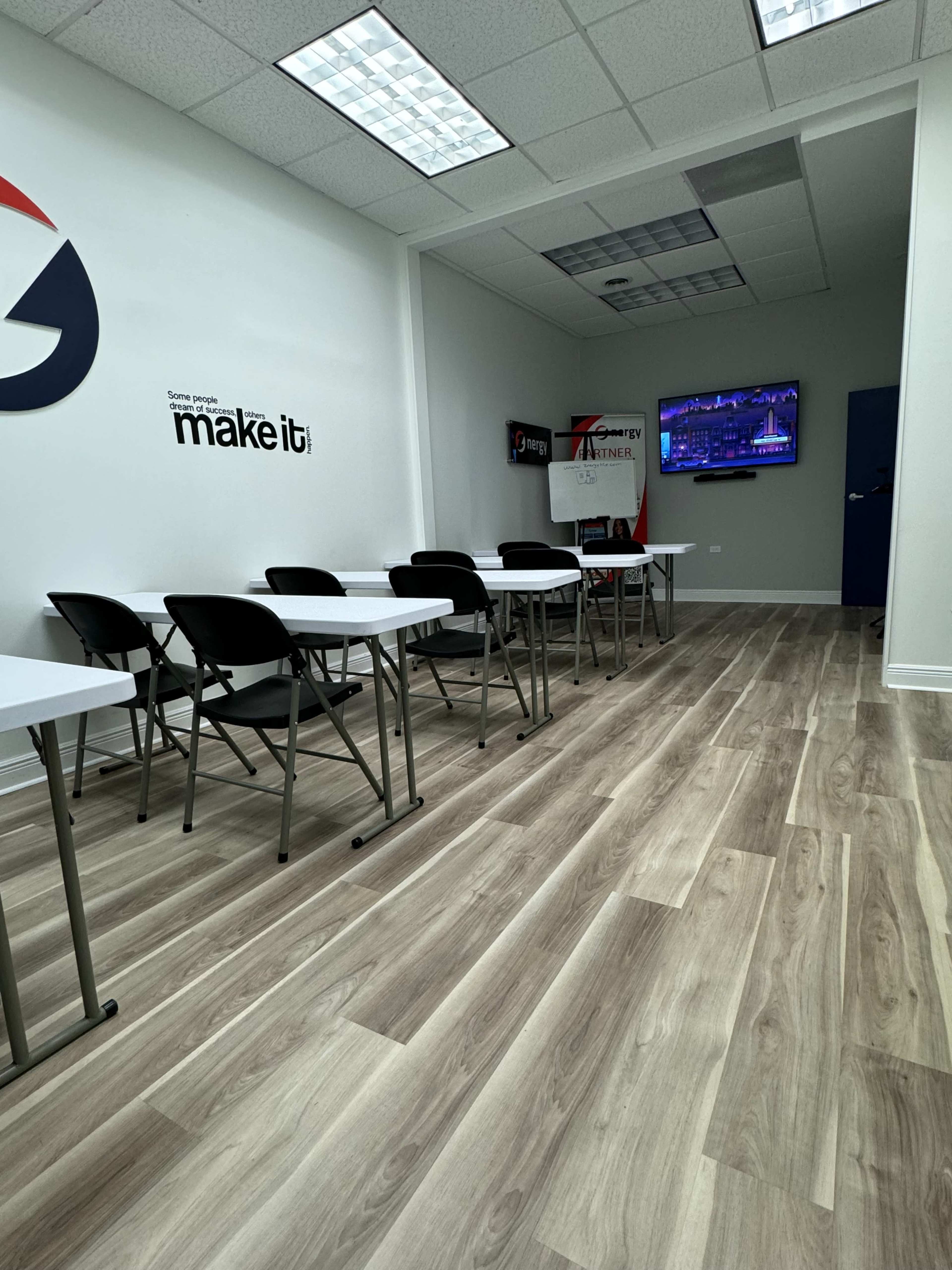 The image shows a classroom setting with rows of white tables and chairs facing a wall-mounted TV in a well-lit room.
