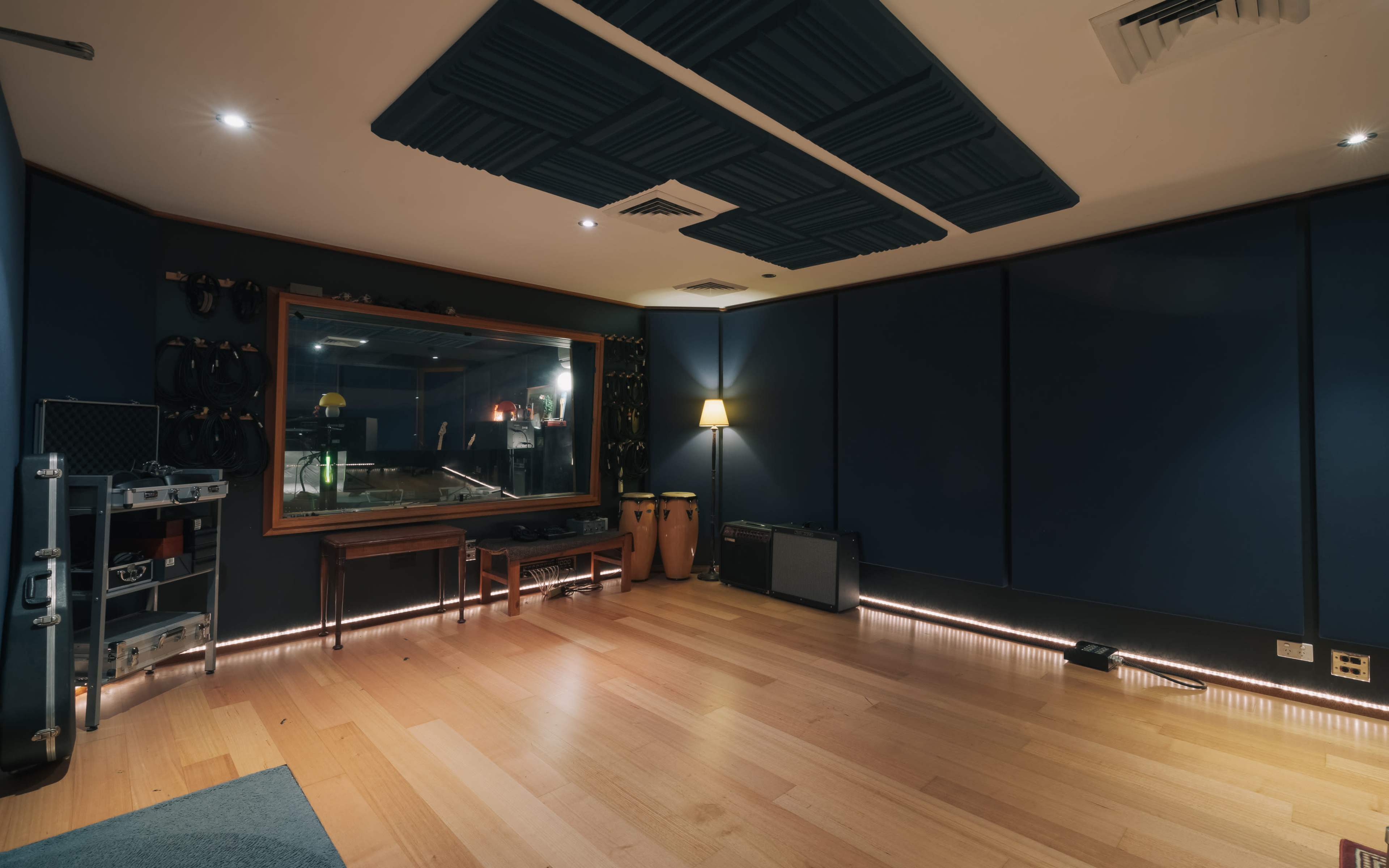 A recording studio with wooden flooring, blue soundproof walls, and a window revealing equipment in a separate room.