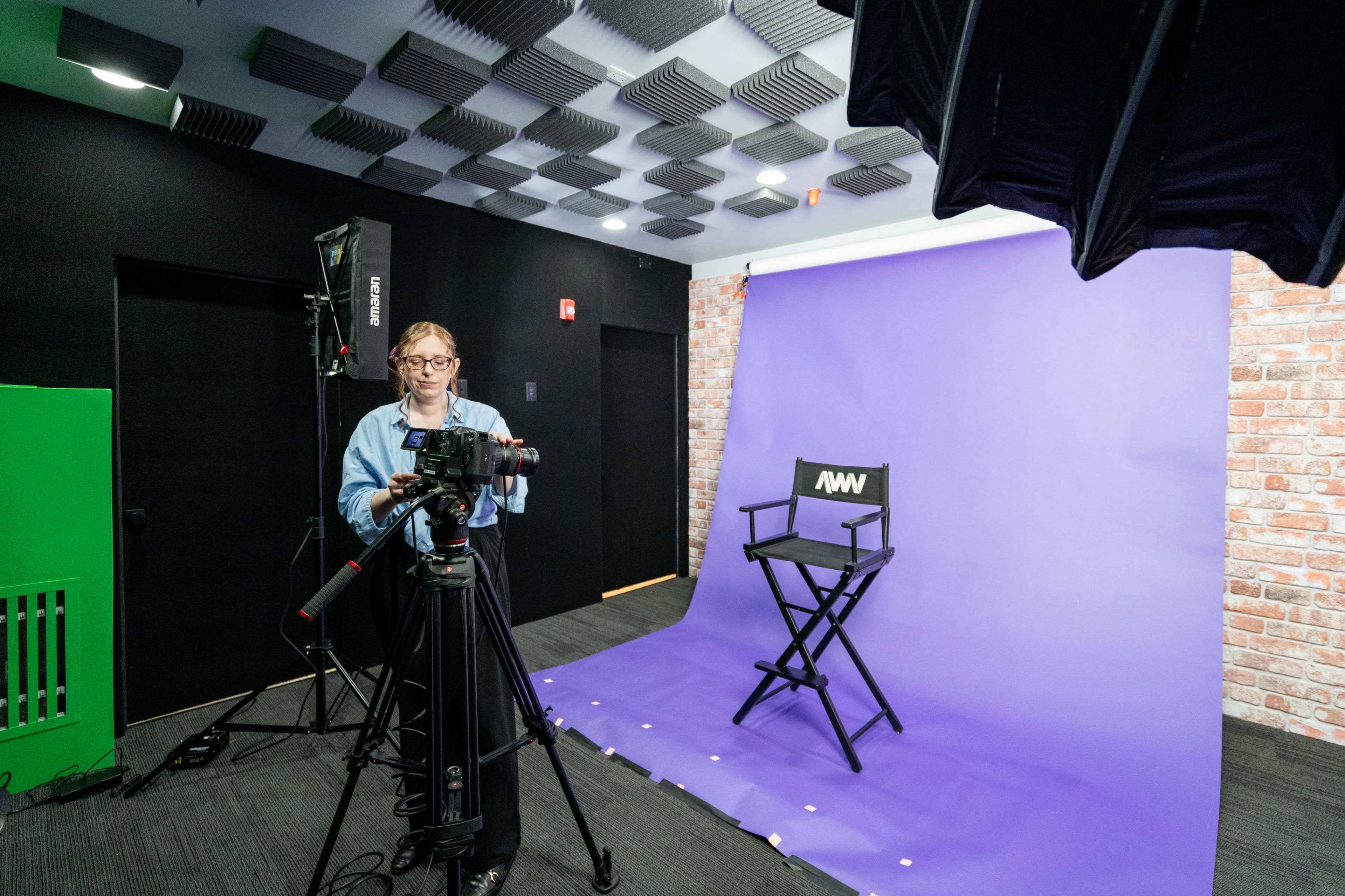 A woman operates a camera in a studio setting with a purple backdrop and a black director's chair.