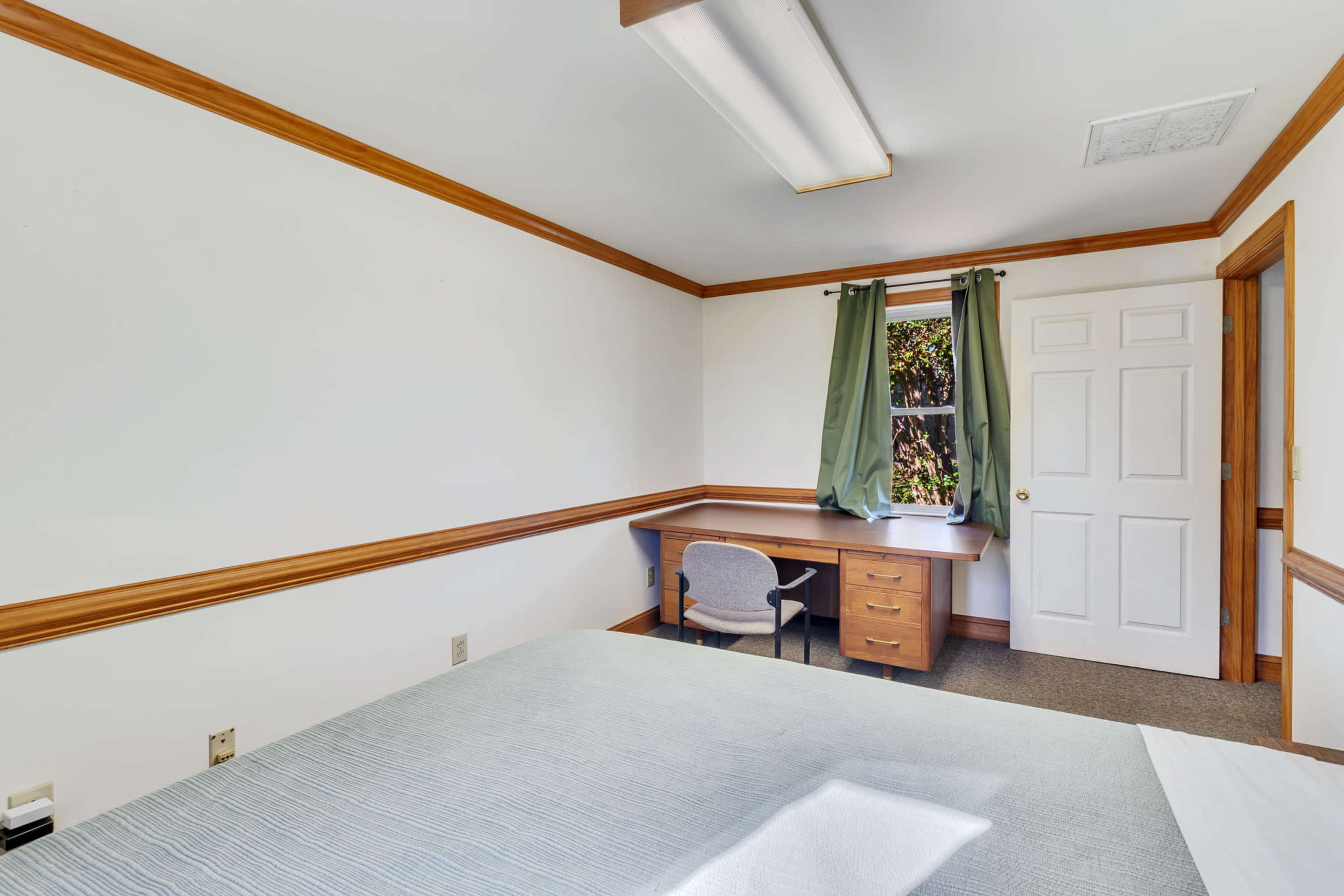 The image shows a simple bedroom featuring a bed, a desk with a chair, and a window with green curtains.
