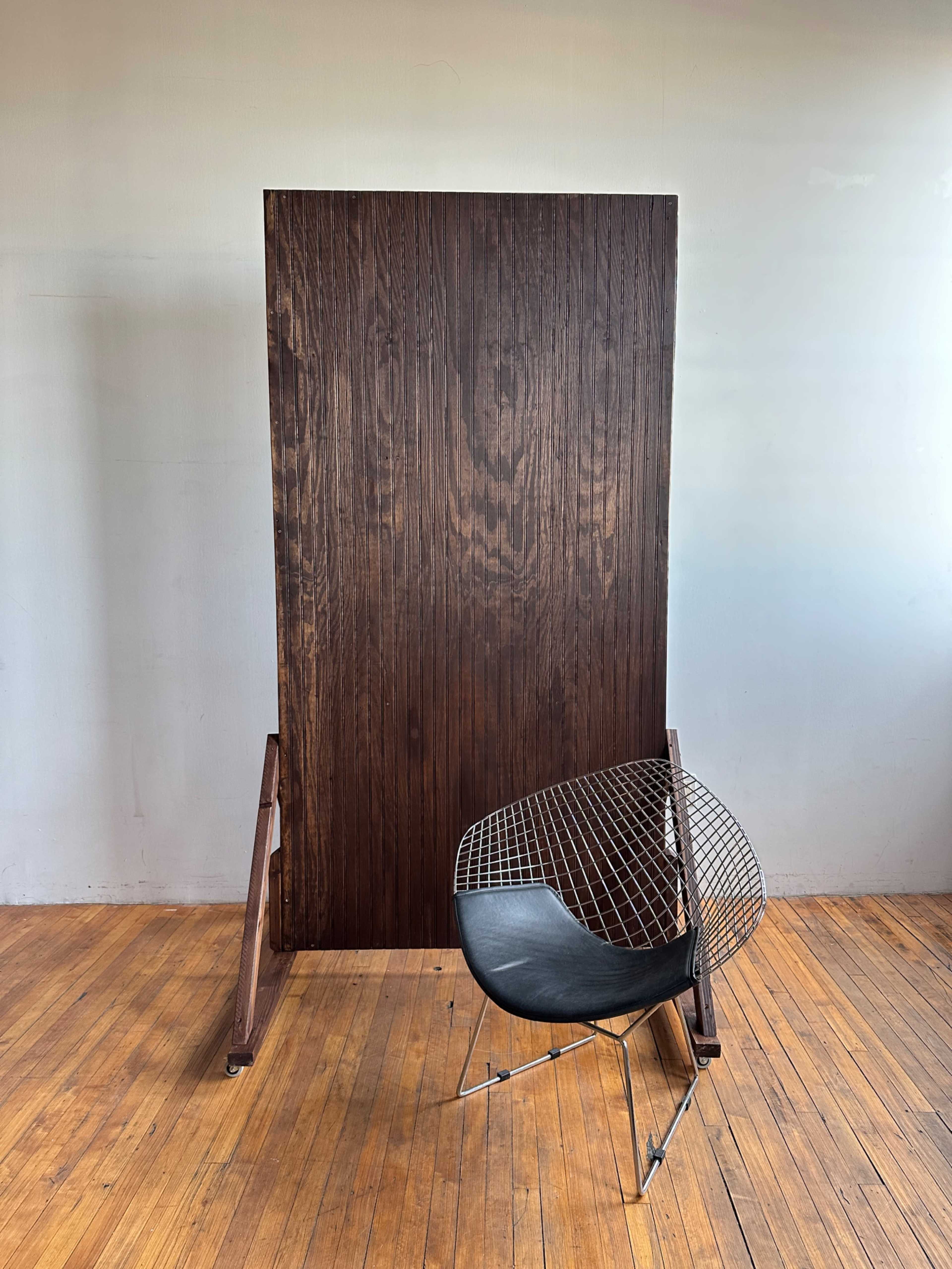 A large wooden panel stands vertically against a wall beside a wire mesh chair on a wooden floor.