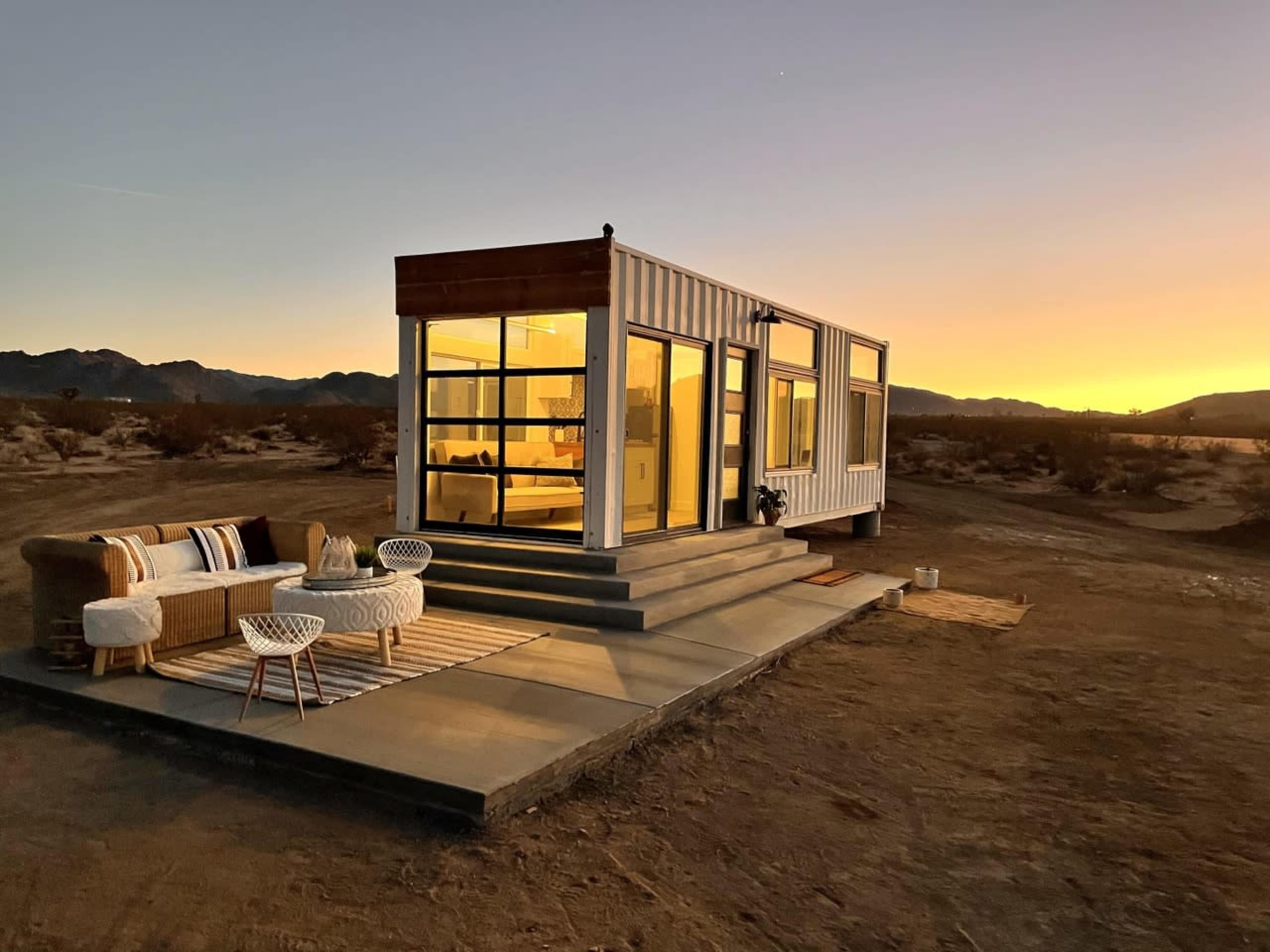 A modern, container-style home with large windows is situated on a desert landscape at sunset, accompanied by outdoor seating.