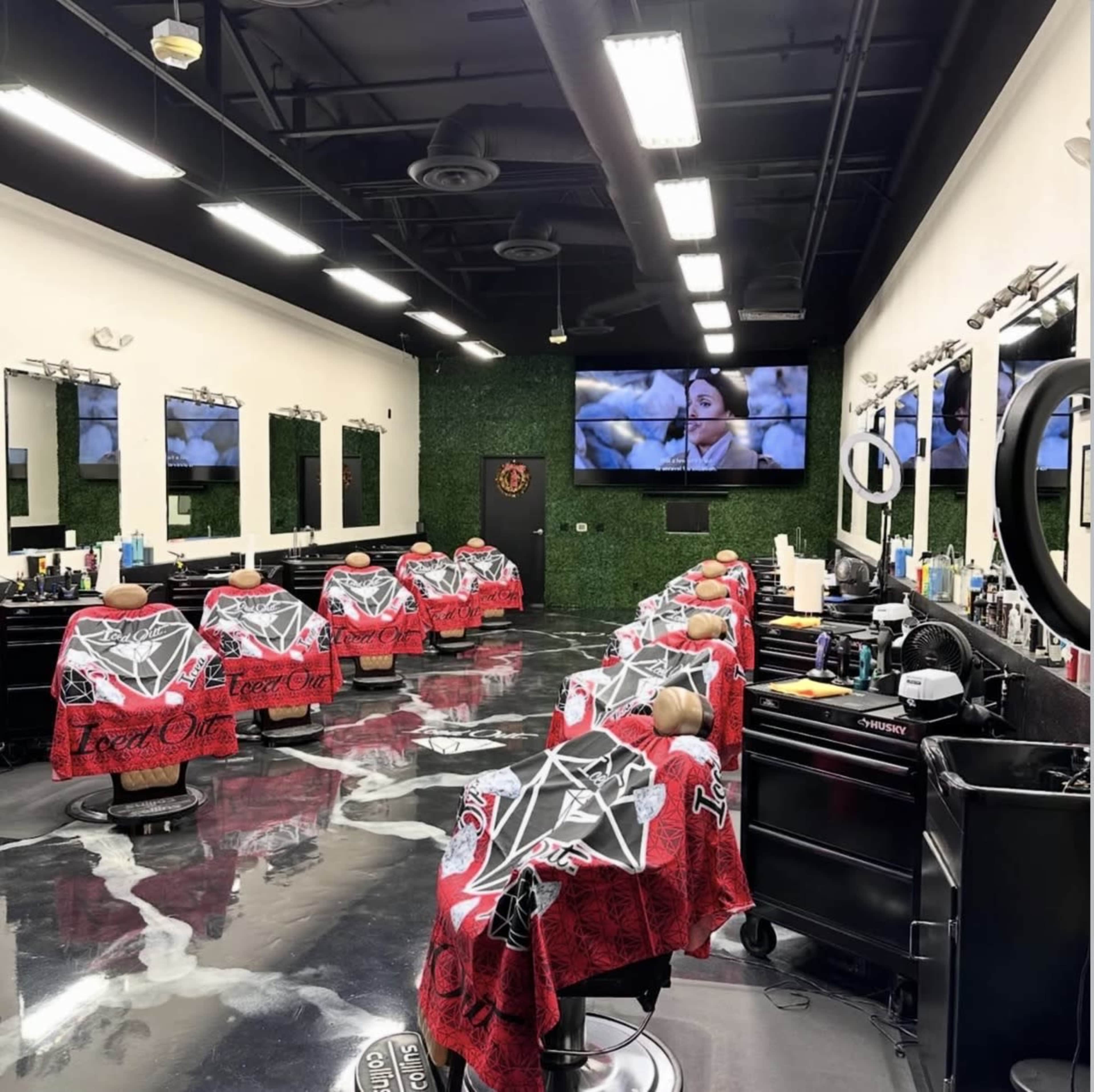 The image shows a modern barbershop with several barber chairs covered in red capes, large mirrors, and a television screen mounted on the wall.