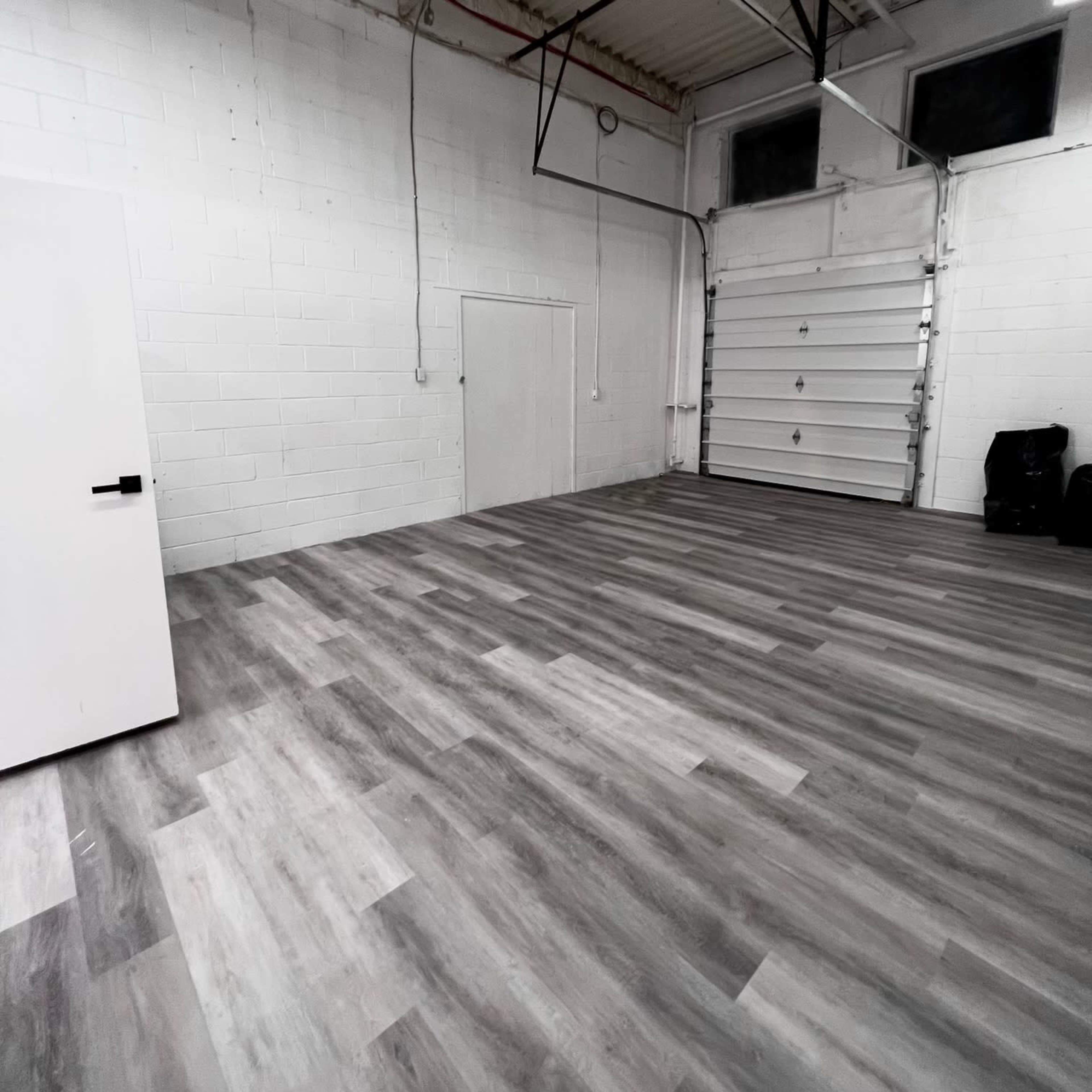 An empty industrial space with gray wooden flooring and a white wall with a door and a large overhead garage door.