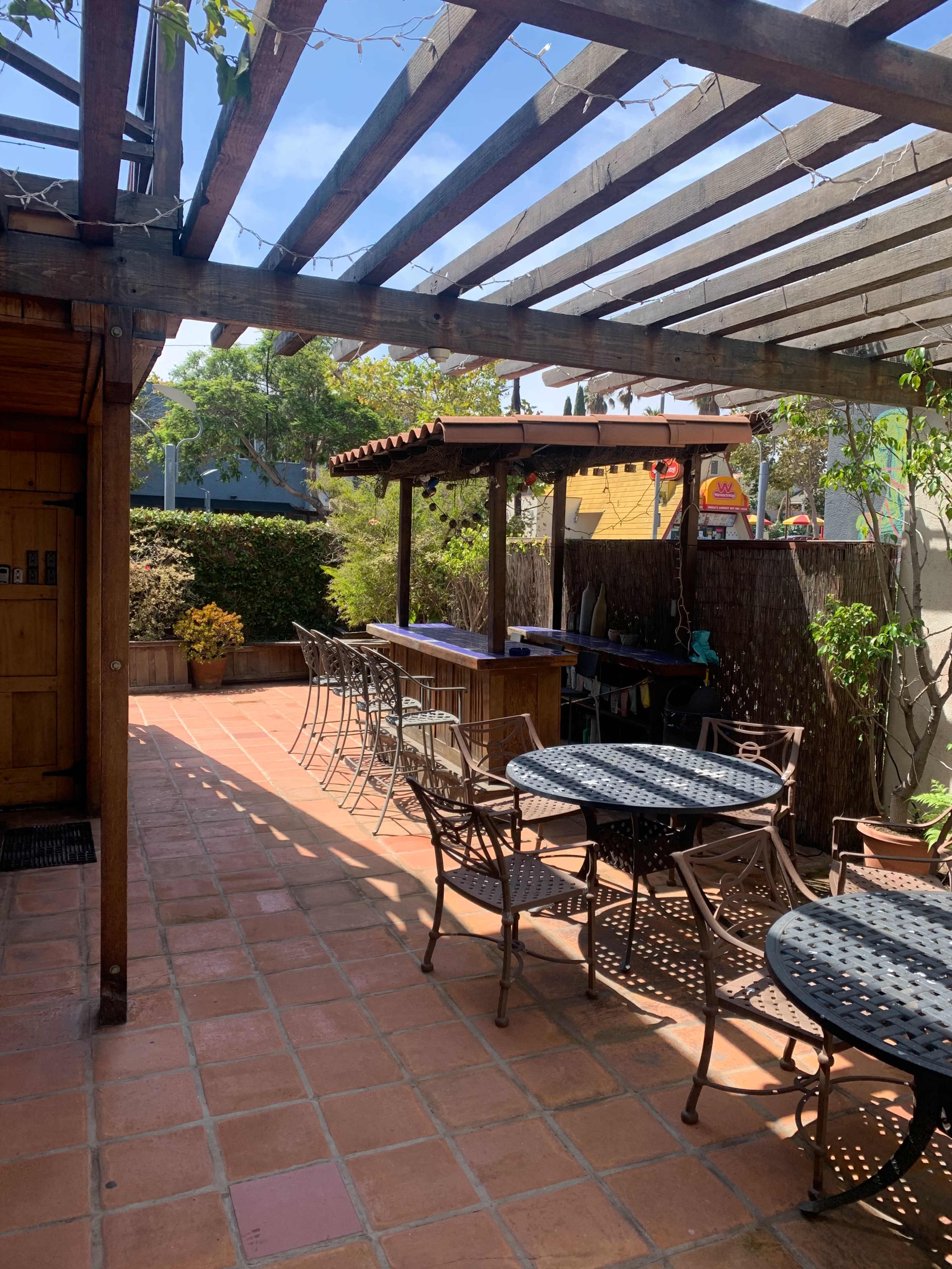 A shaded outdoor patio features wooden pergolas, metal tables and chairs, and potted plants on a terracotta tiled floor.