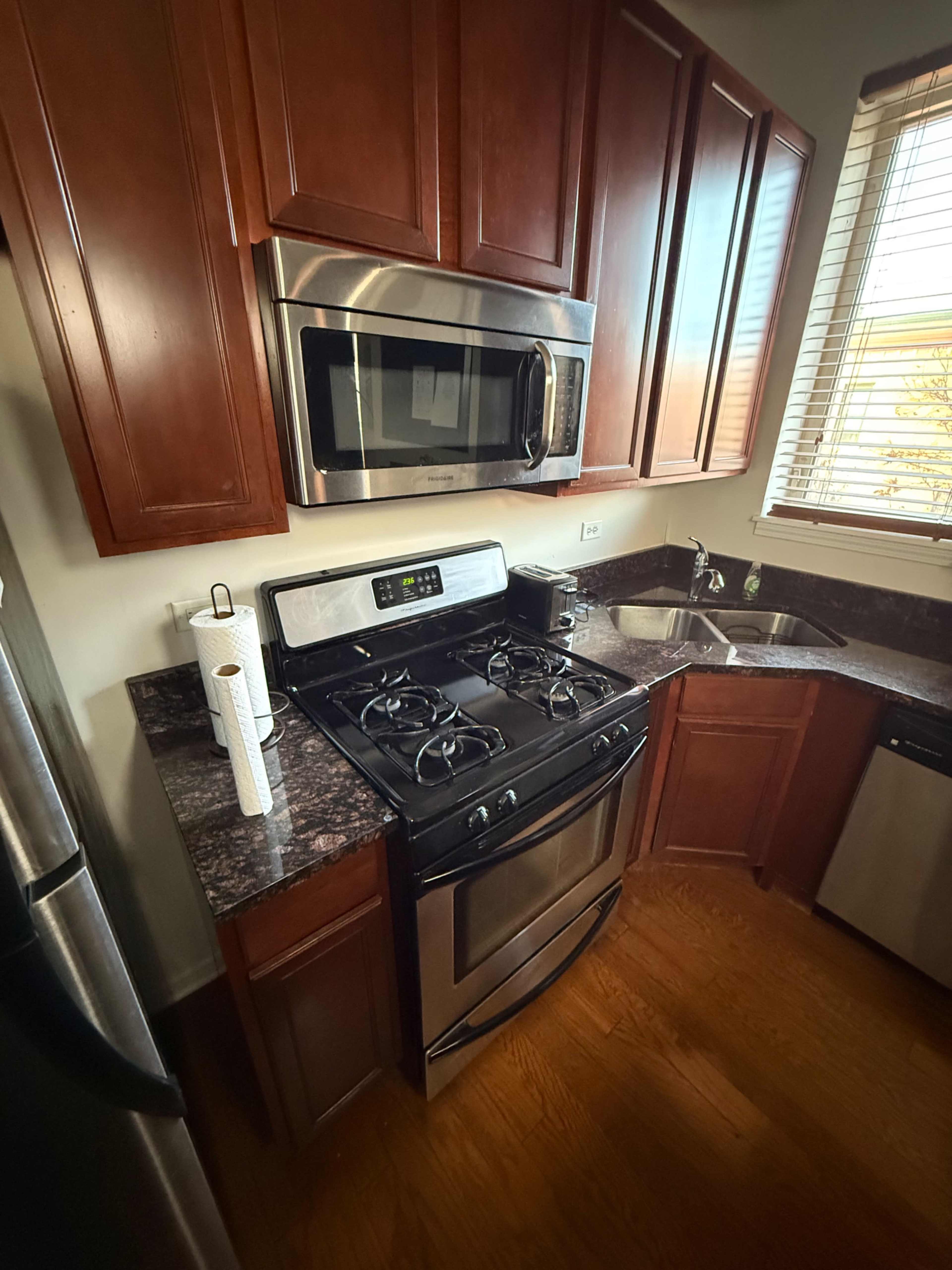 The image shows a compact kitchen featuring dark wooden cabinetry, a stainless steel microwave above a gas stove, and a sink with a window above it.