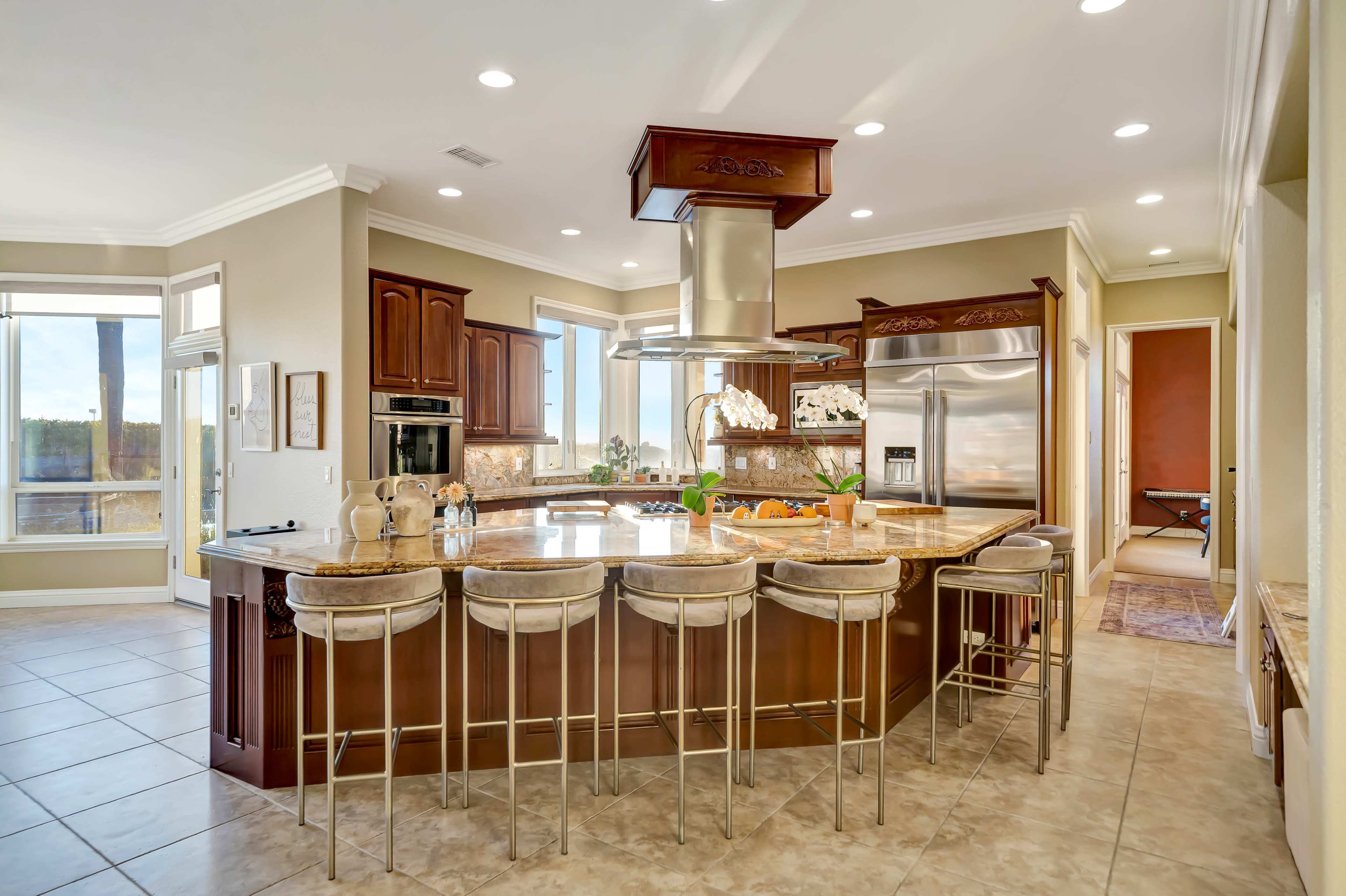 The image shows a modern kitchen with wooden cabinetry, a central island with bar seating, and stainless steel appliances.