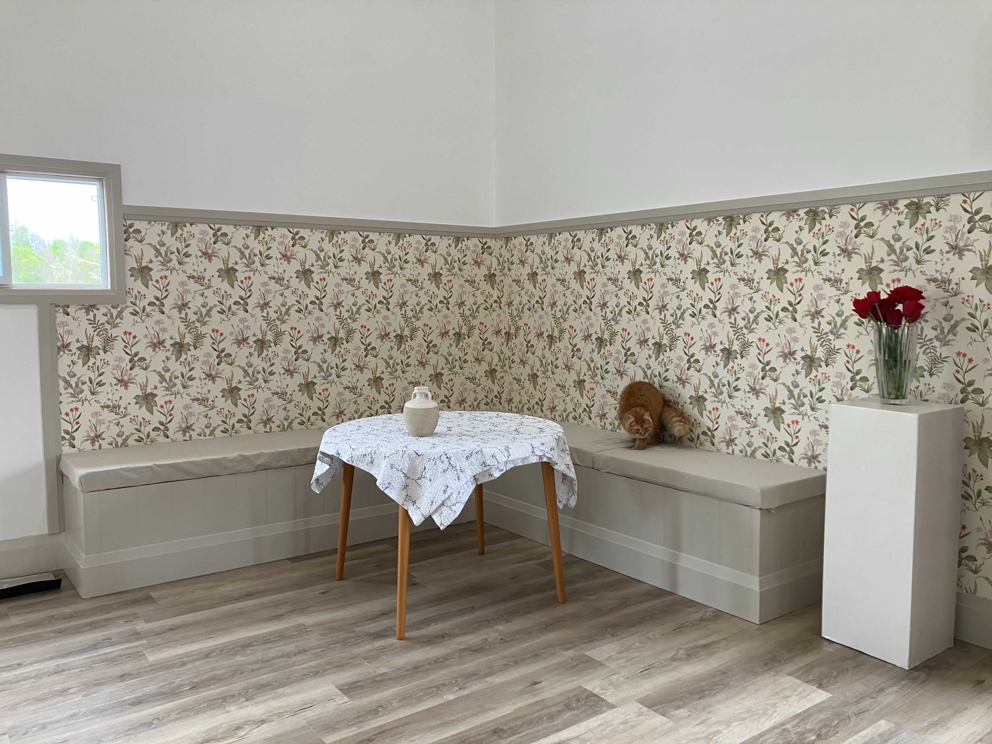 The image shows a corner of a room with a floral wallpaper, a round table covered with a tablecloth, a decorative vase with flowers, and a cat resting on a bench.