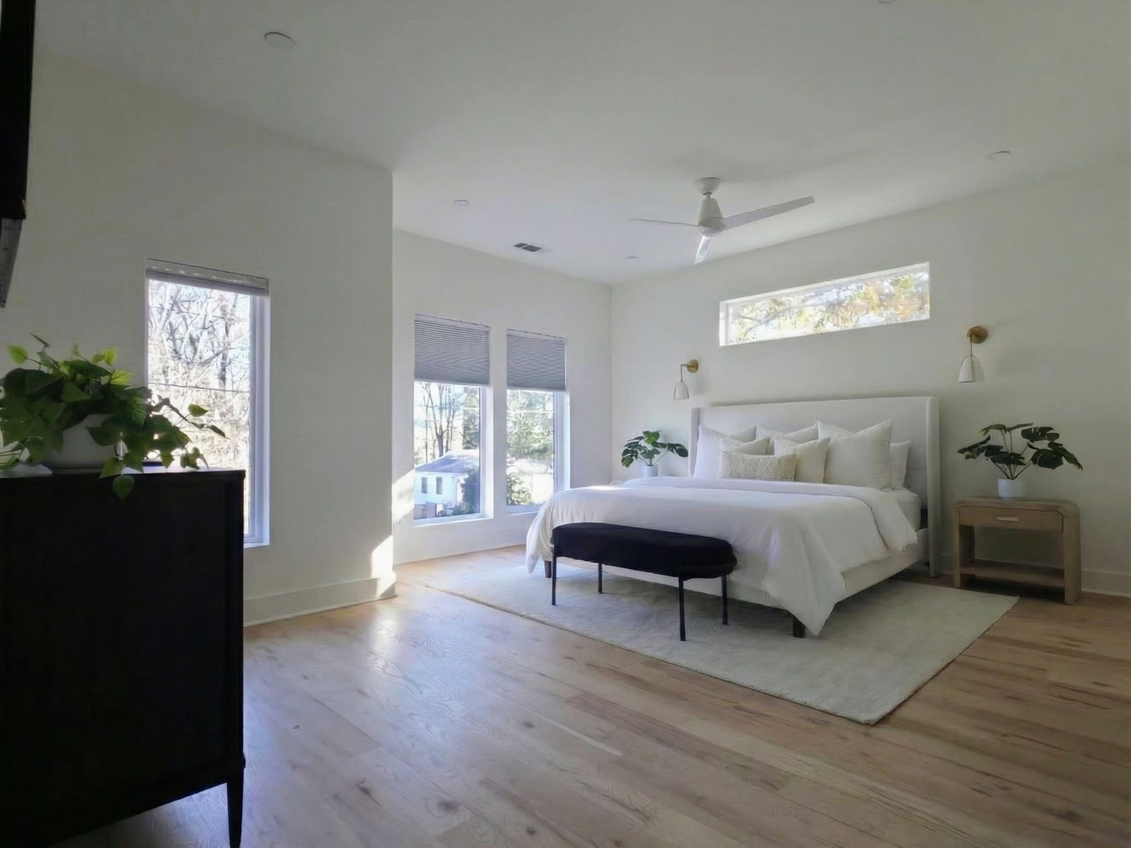 A spacious bedroom features a large bed with white bedding, a bench at the foot, and large windows letting in natural light.