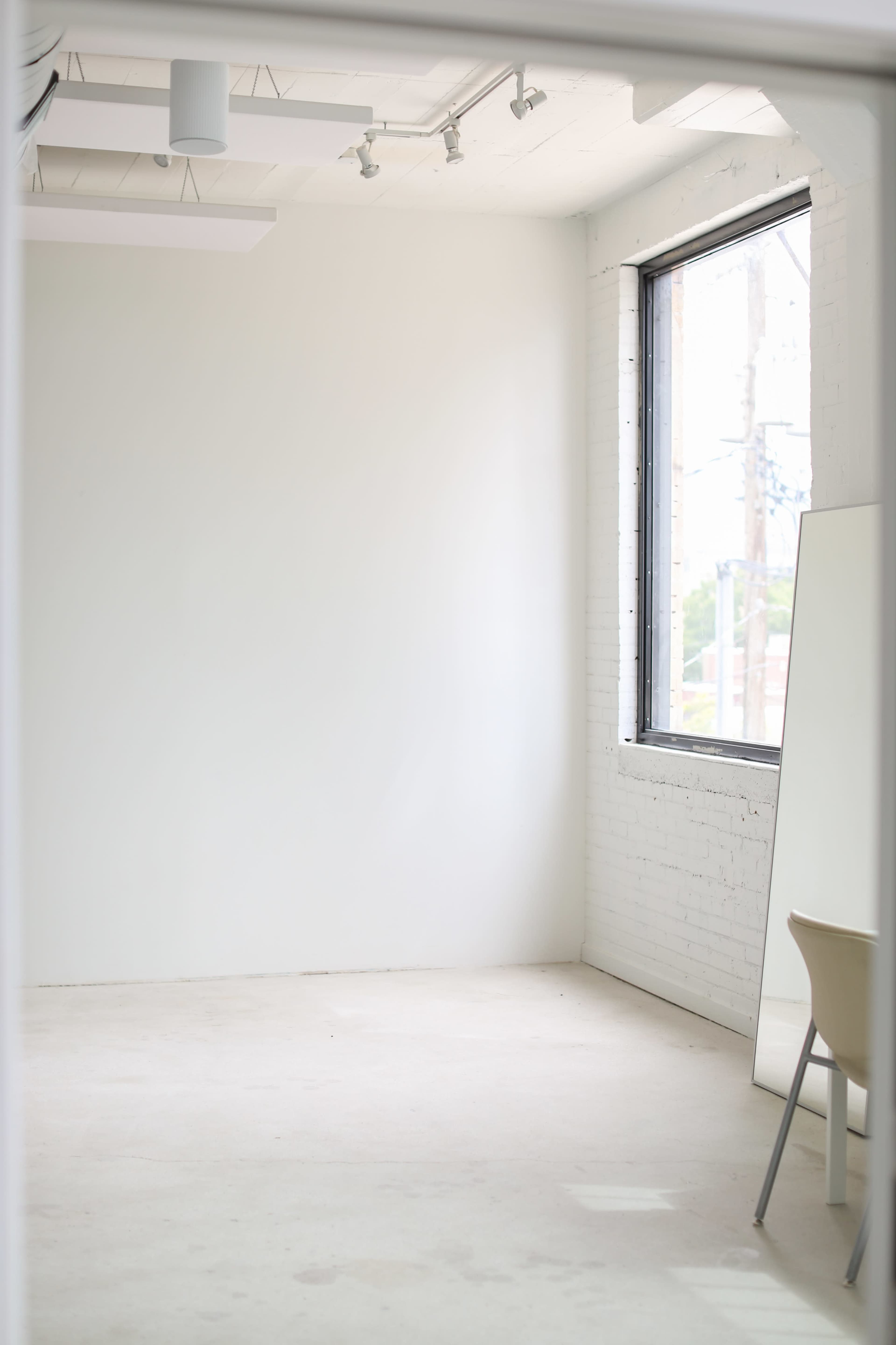 An empty, well-lit room with white walls, a large window, and a chair next to a mirror.