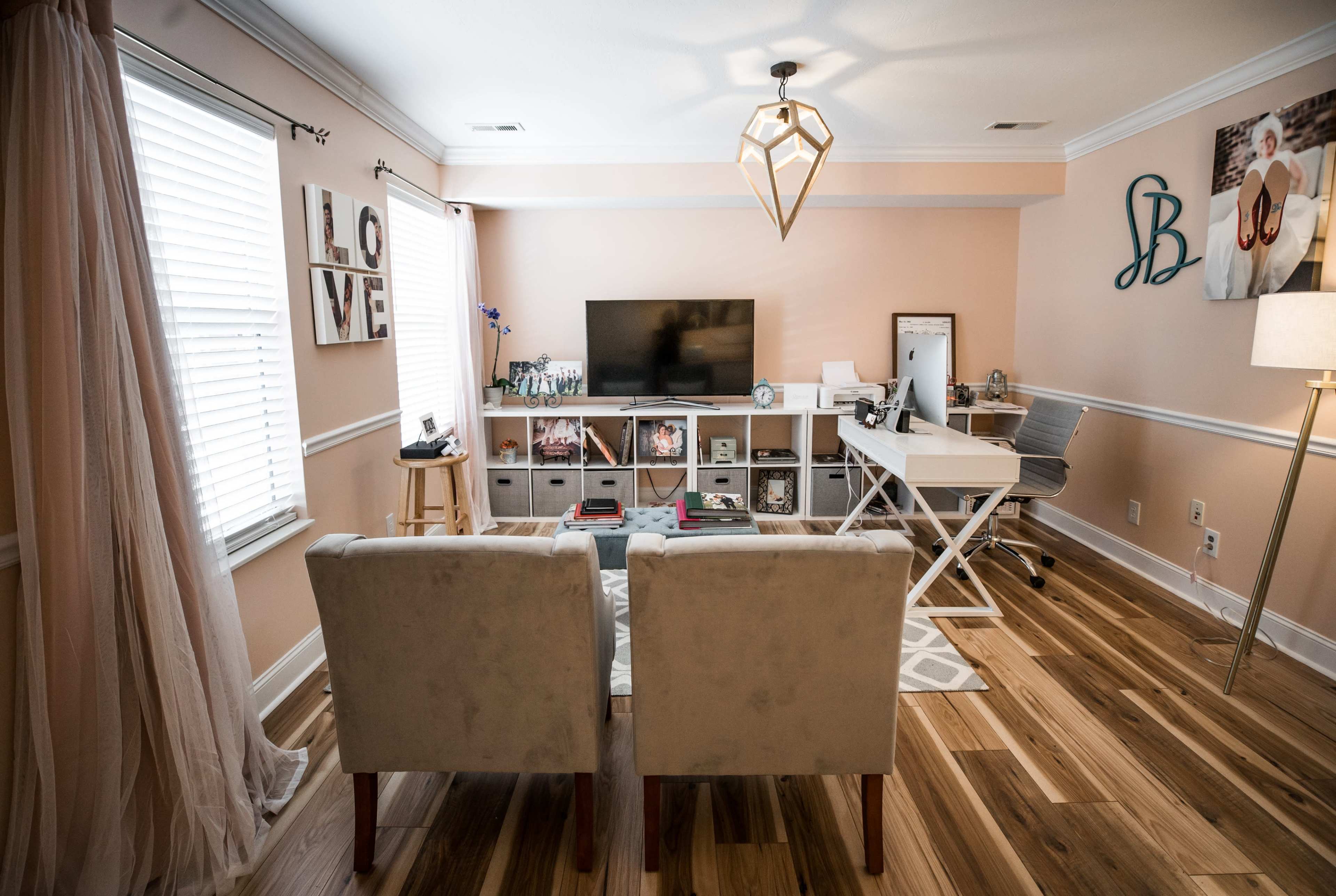 A modern home office features a light-colored sofa, a desk facing a television, and wooden flooring, with decorative items and plants in the background.