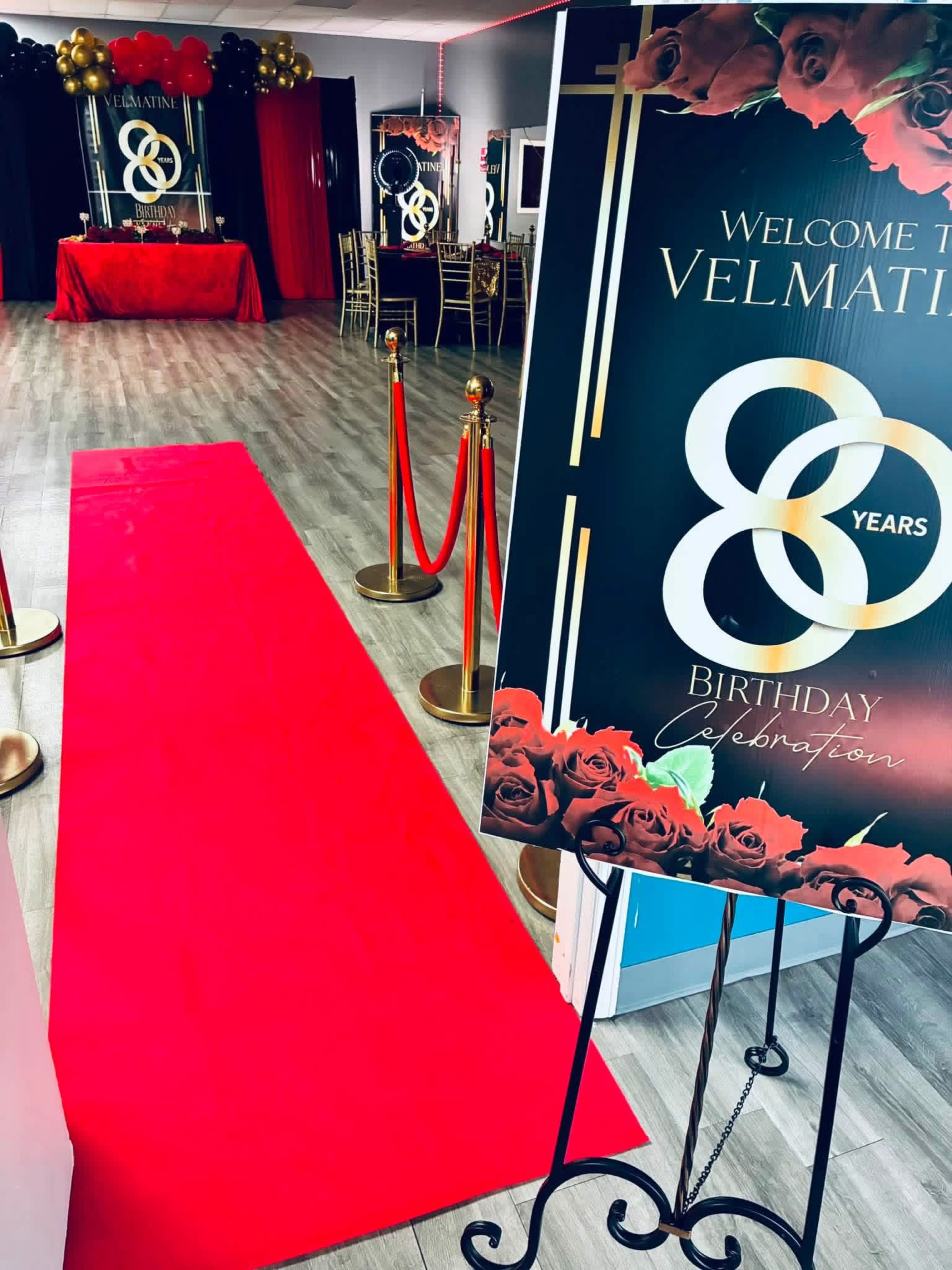 The image shows a banquet hall decorated for an 80th birthday celebration, featuring a red carpet entrance and a welcome sign adorned with roses.
