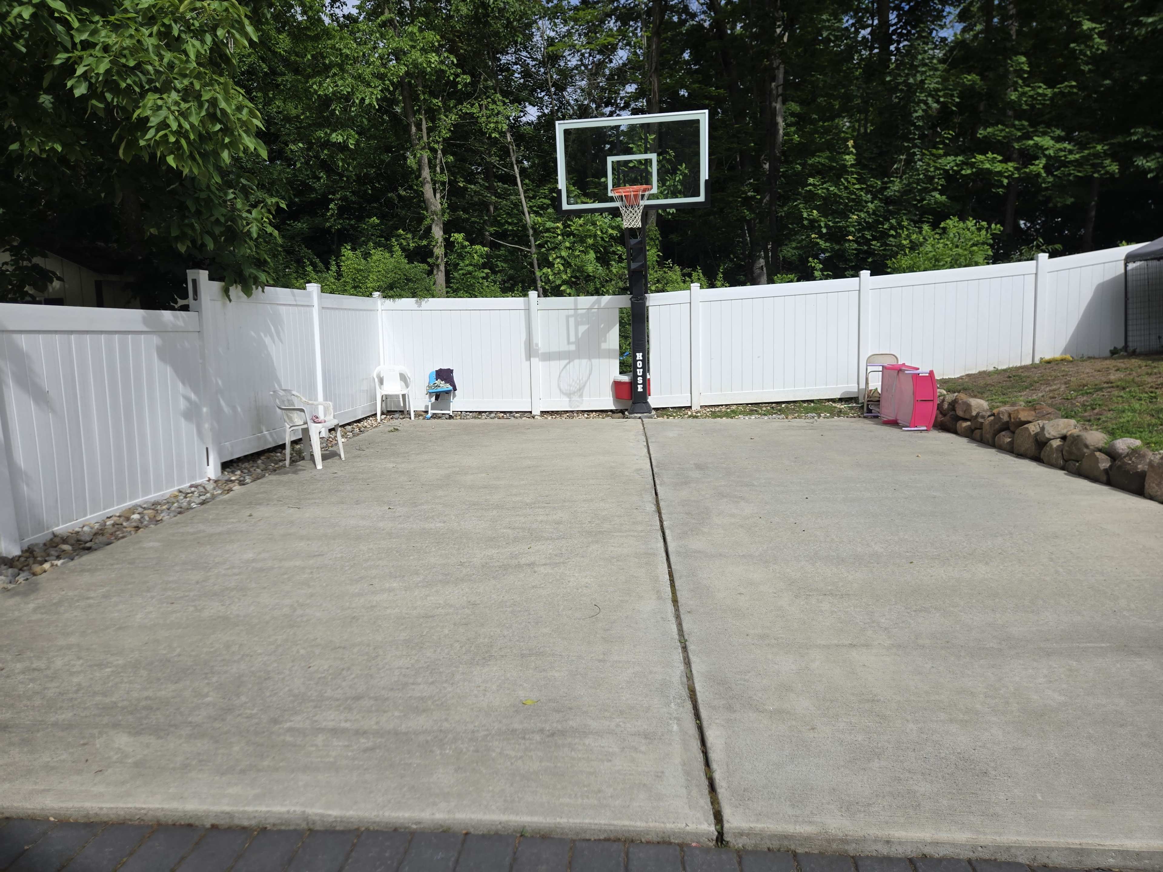 The image shows a concrete outdoor space with a basketball hoop, surrounded by a white fence and greenery in the background.