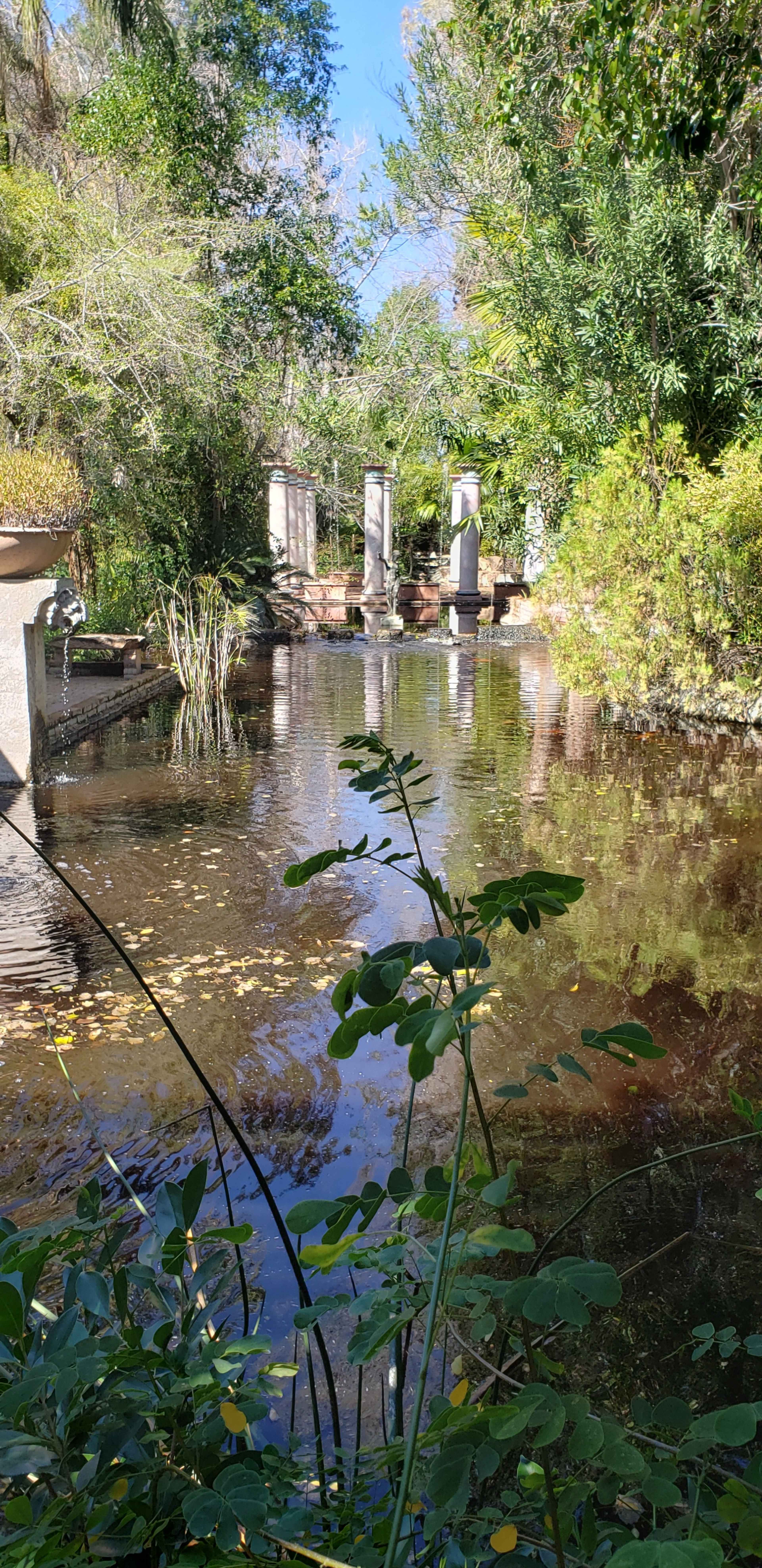 A calm pond is surrounded by lush greenery and features stone pillars in the background.
