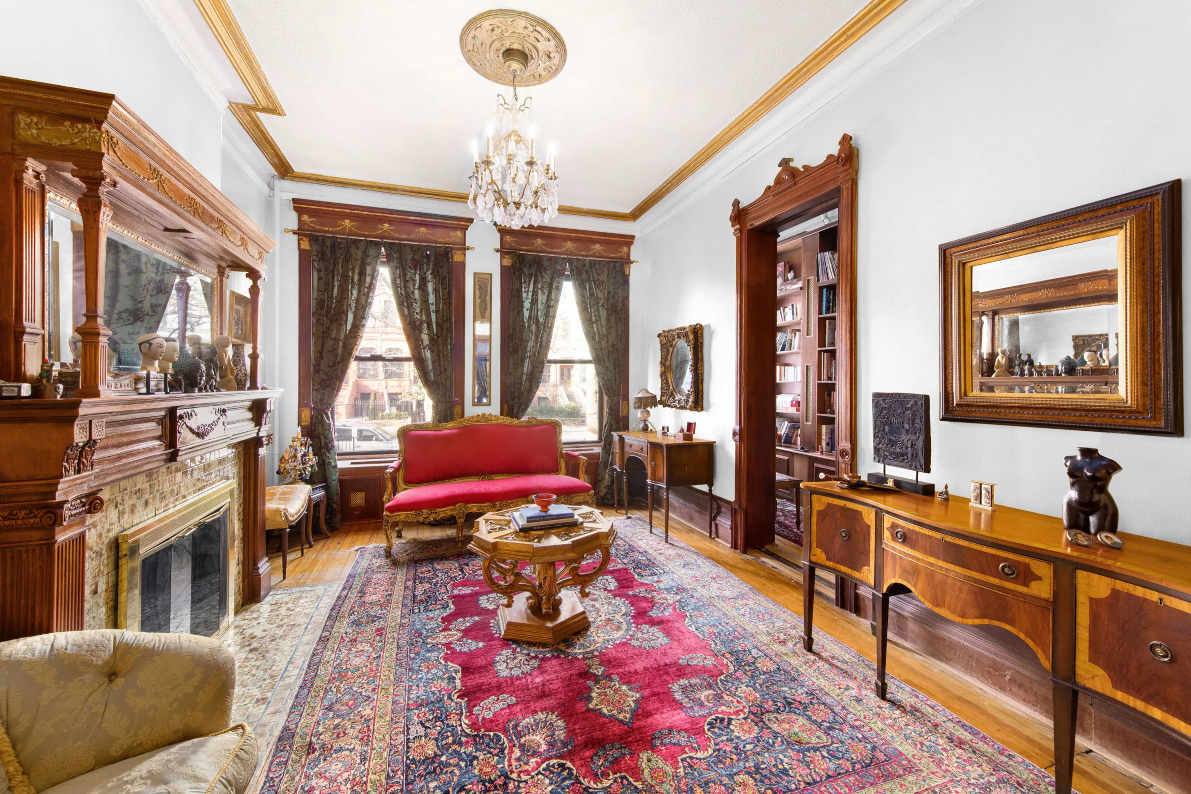 A vintage living room featuring ornate wood furniture, a patterned rug, a chandelier, and large windows with dark curtains.
