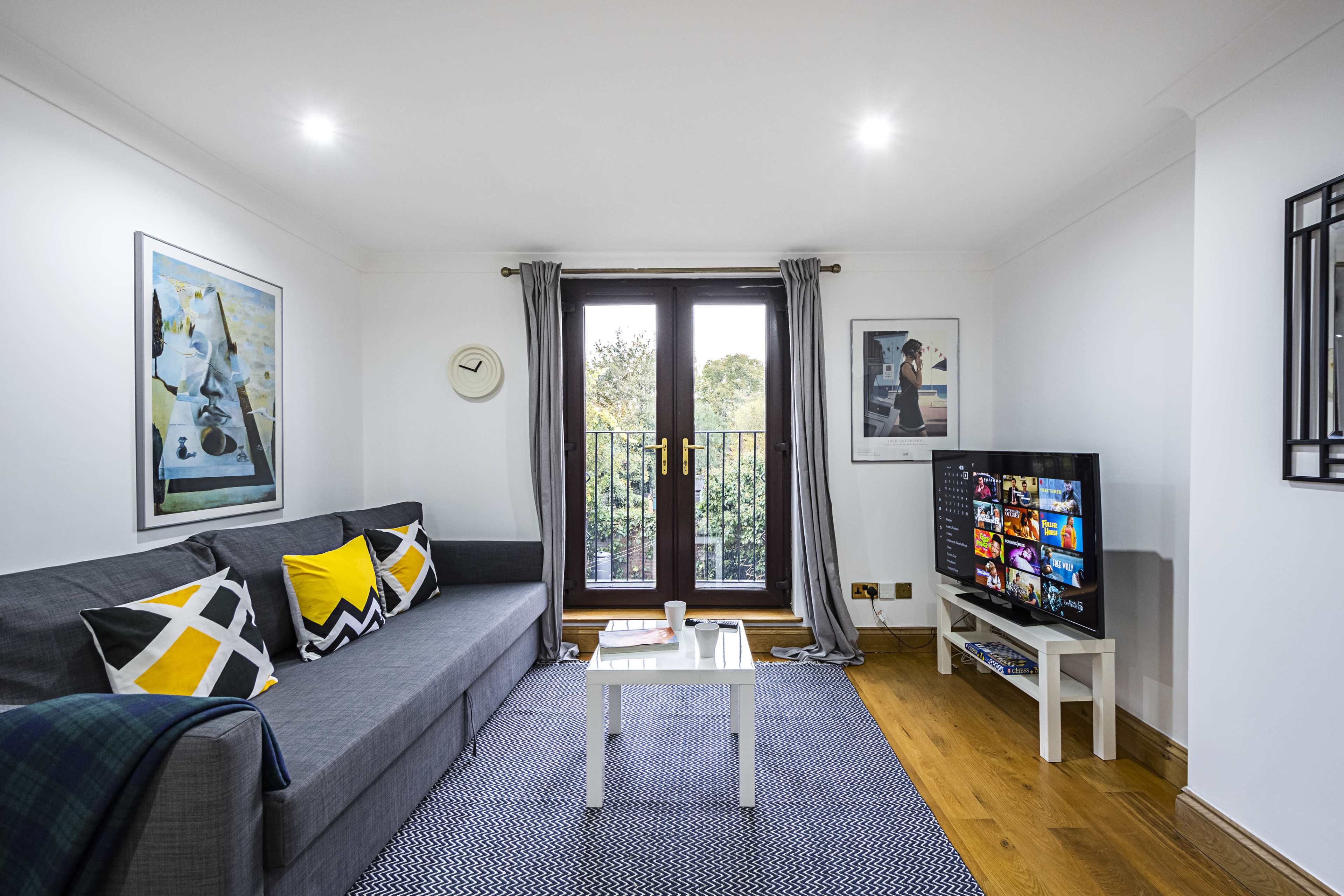 A modern living room features a gray sofa with patterned cushions, a wall-mounted TV, and large windows opening to a balcony.