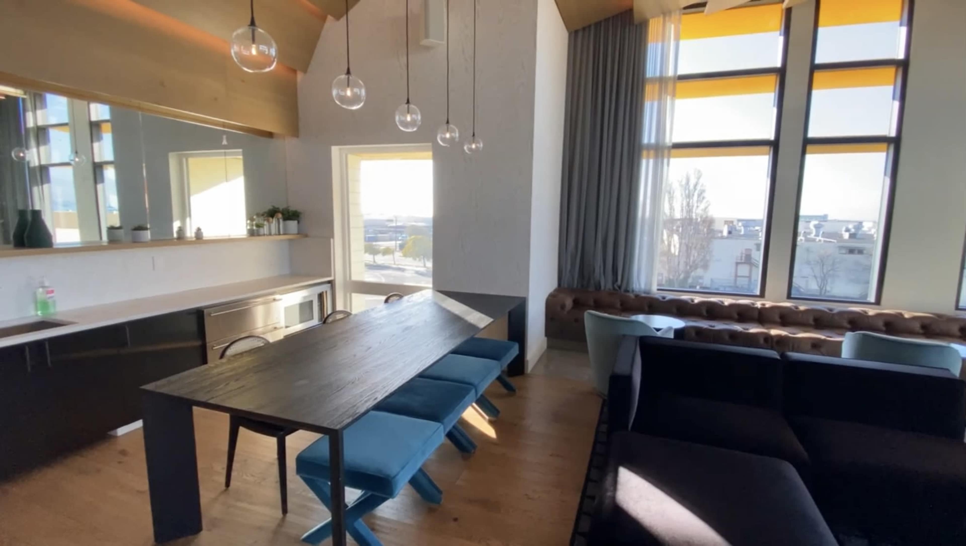 A modern living space featuring a dining table with six blue chairs, a dark sofa, large windows with yellow shades, and a kitchen area in the background.