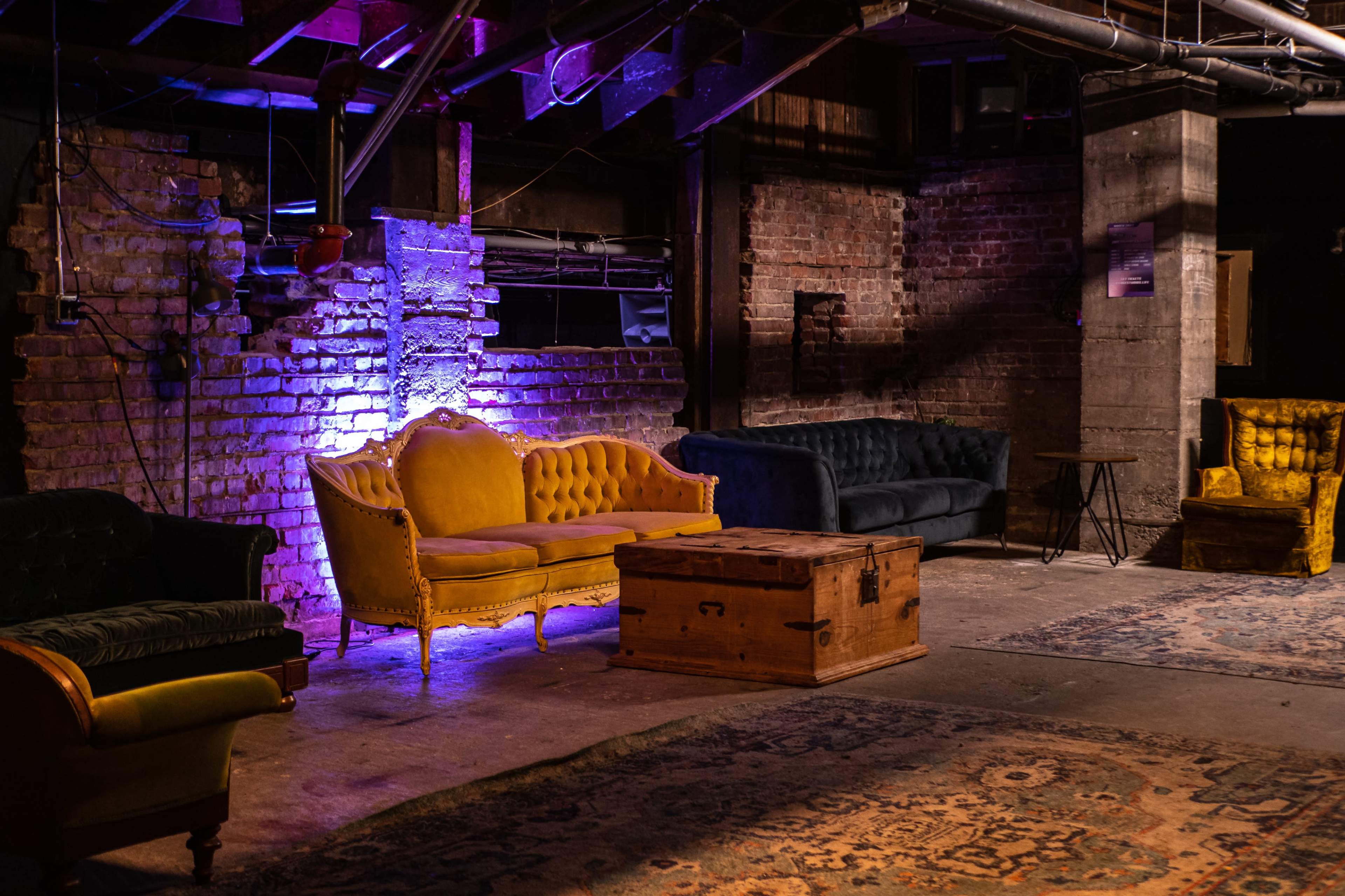 A dimly lit indoor space with various vintage-style sofas, a wooden coffee table, and area rugs against a backdrop of exposed brick walls.