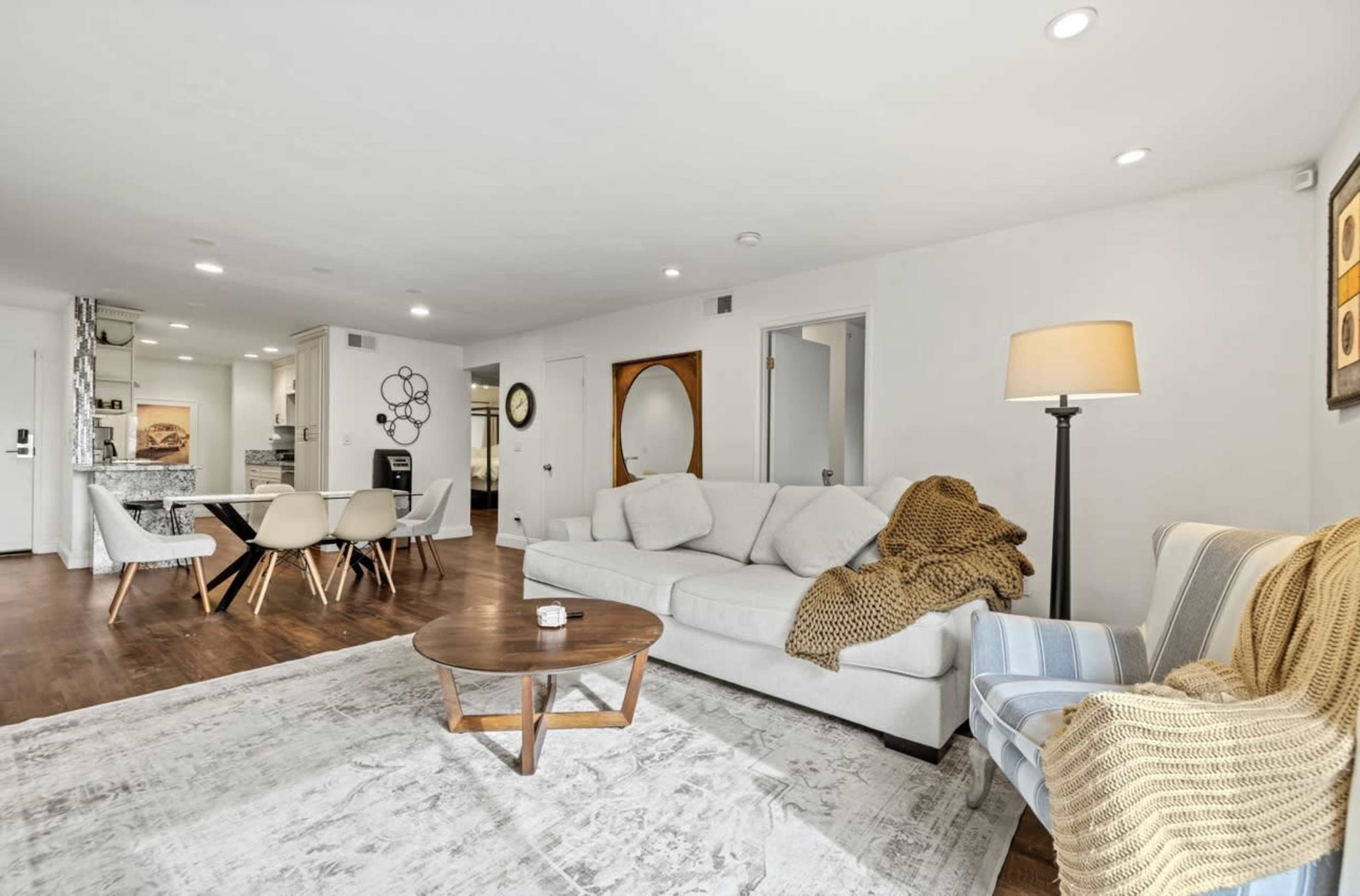 The image shows a living room with a light-colored sofa, a round coffee table, and a patterned rug, with an open kitchen visible in the background.