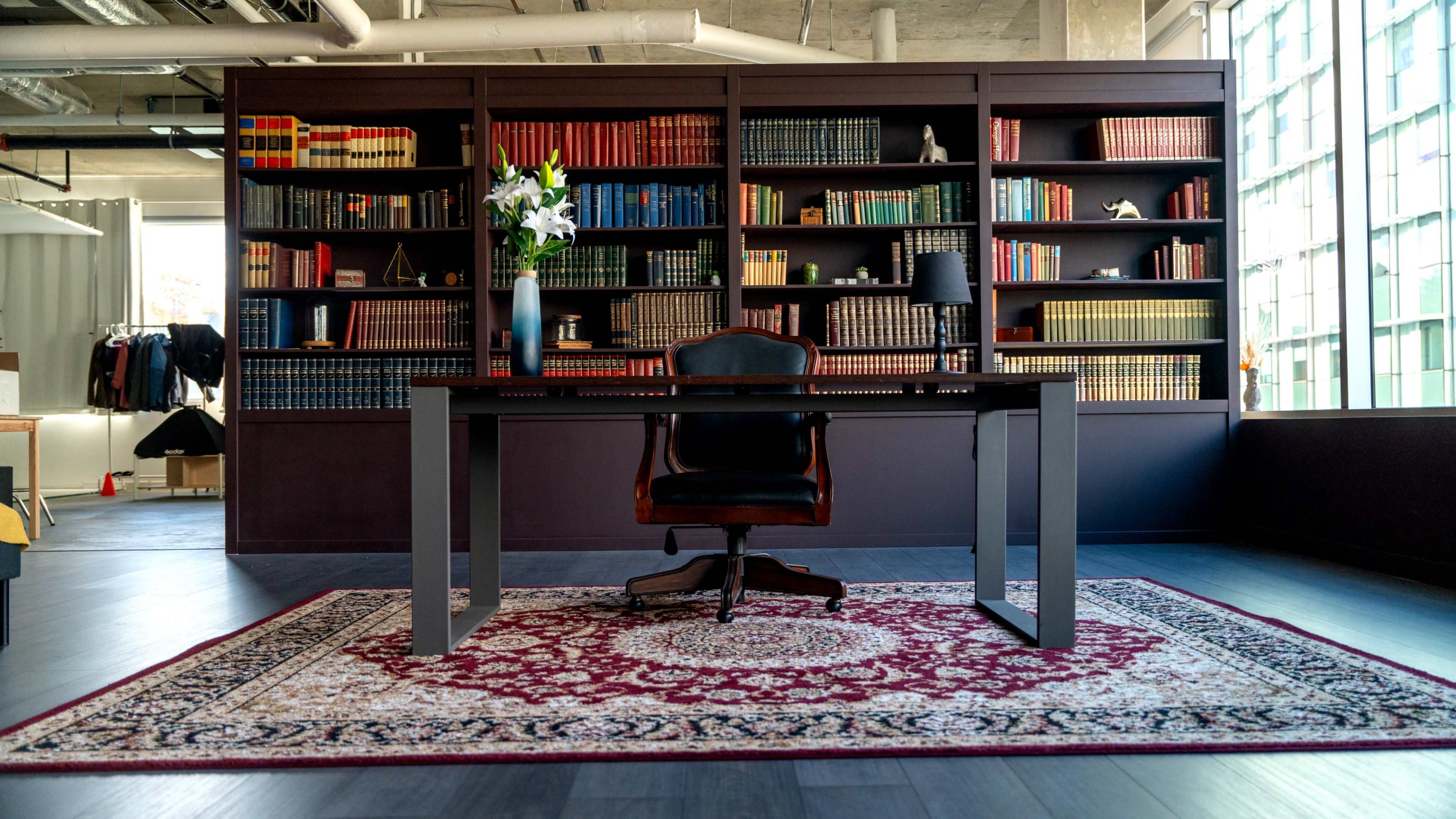 A modern office space features a large desk with a chair in front of a shelf filled with books, and a rug on the floor.