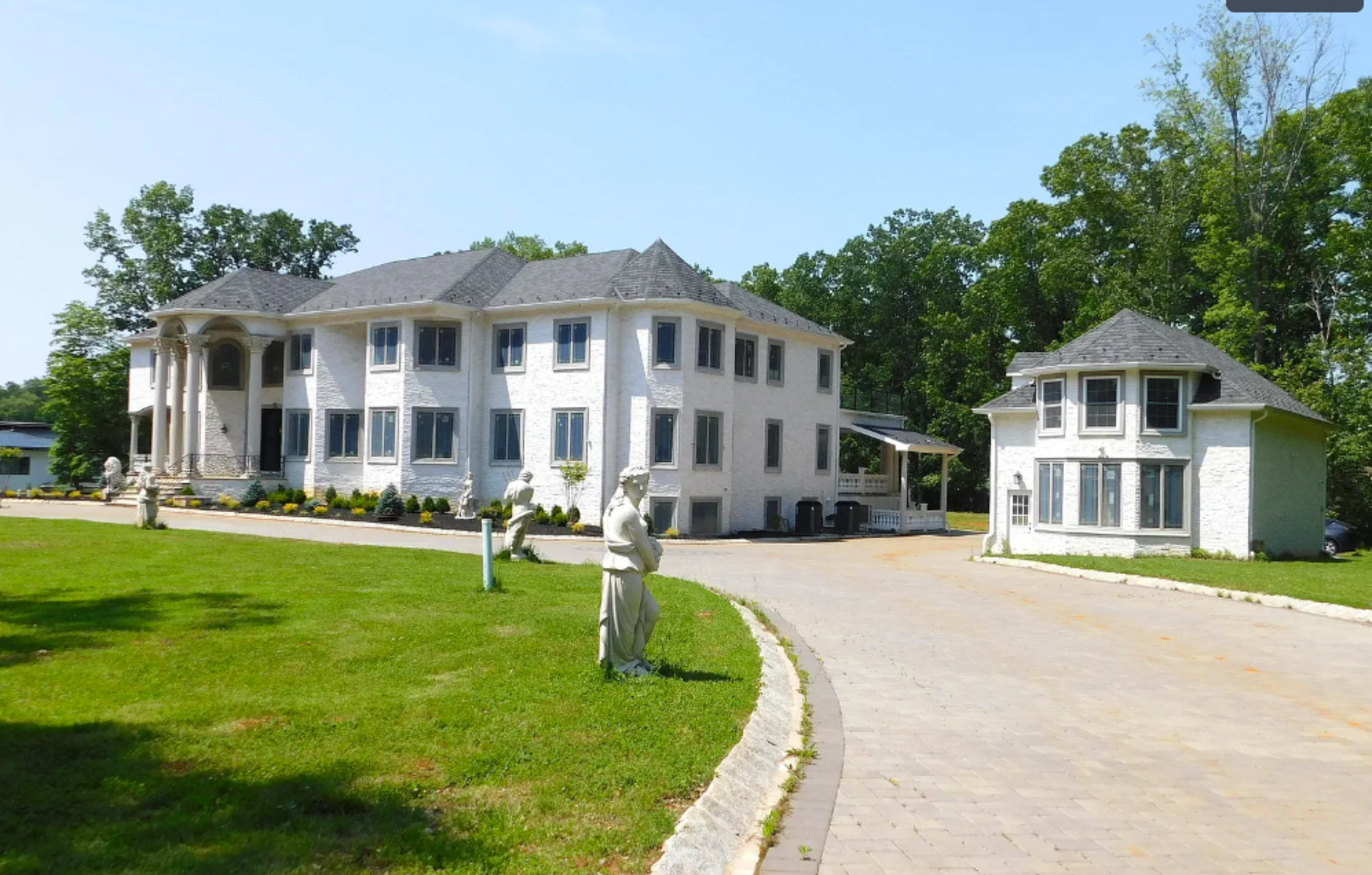 A large white mansion is situated beside a smaller outbuilding, both surrounded by well-maintained landscaping and a paved driveway.