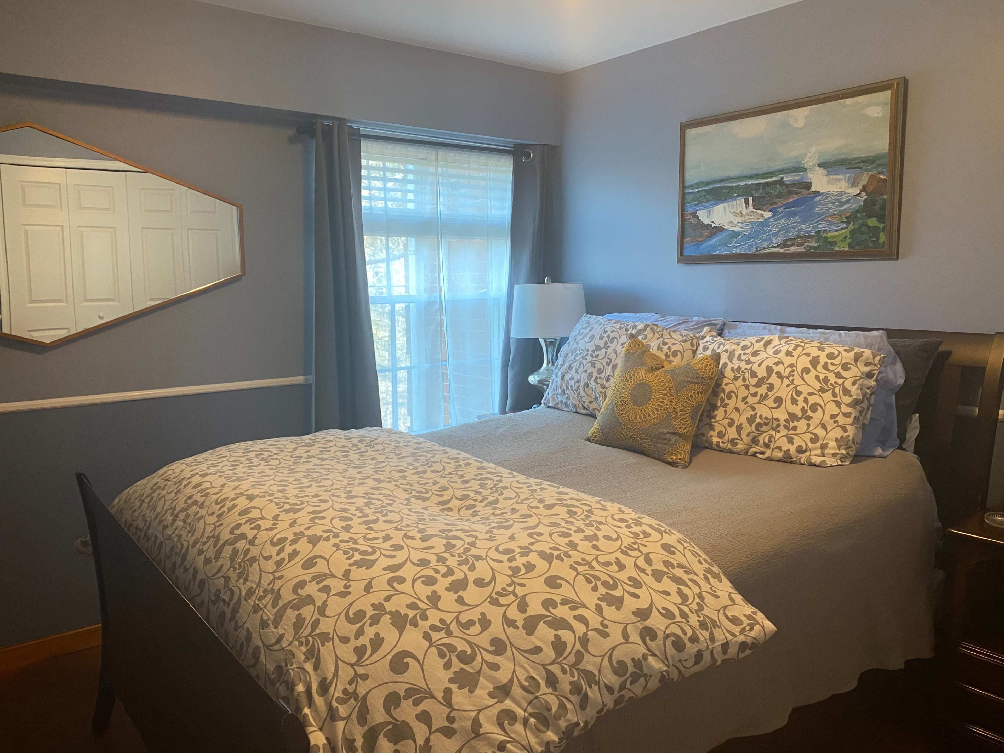 A bedroom with a bed adorned with a patterned comforter, a decorative pillow, a lamp on a nightstand, and artwork on the wall, with a window in the background.
