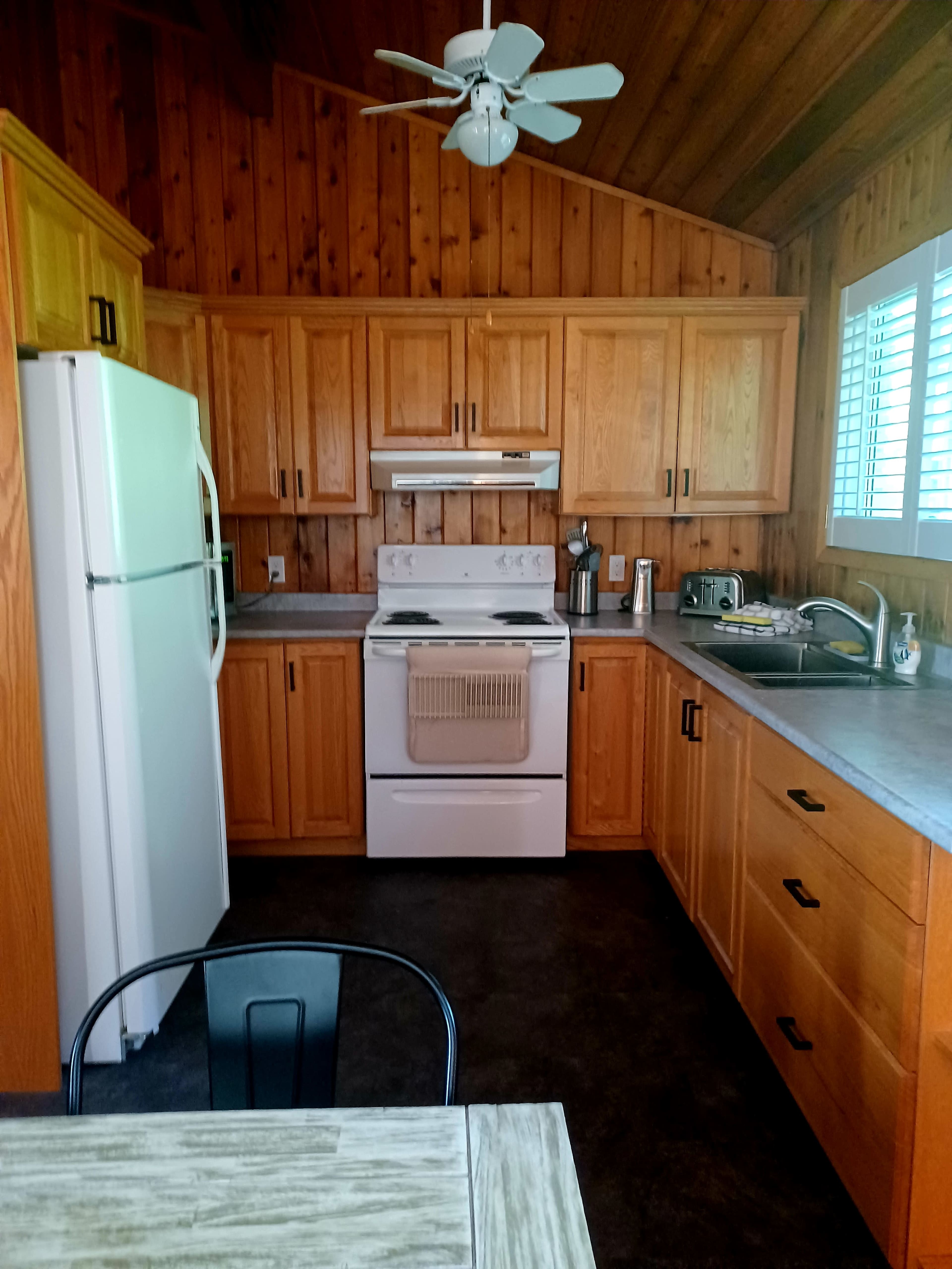 A wood-paneled kitchen with a white refrigerator, stove, and sink, along with wooden cabinets and a small dining table.