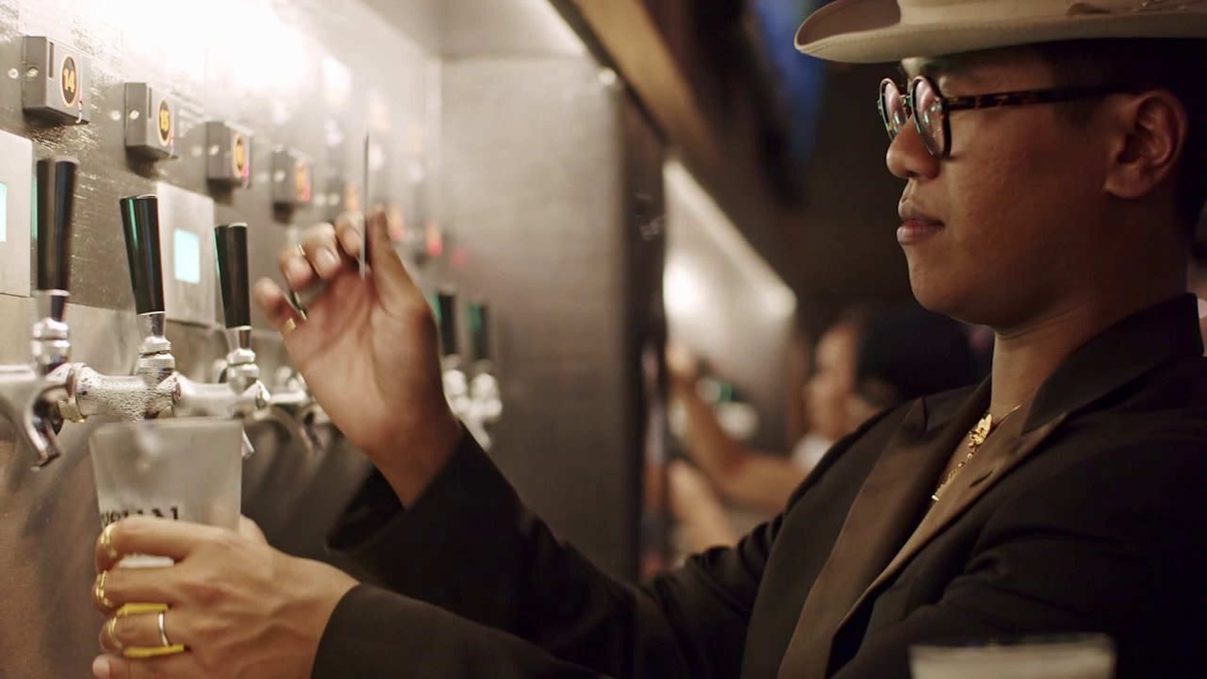 A person wearing a hat and glasses is pouring a drink from a tap at a bar.