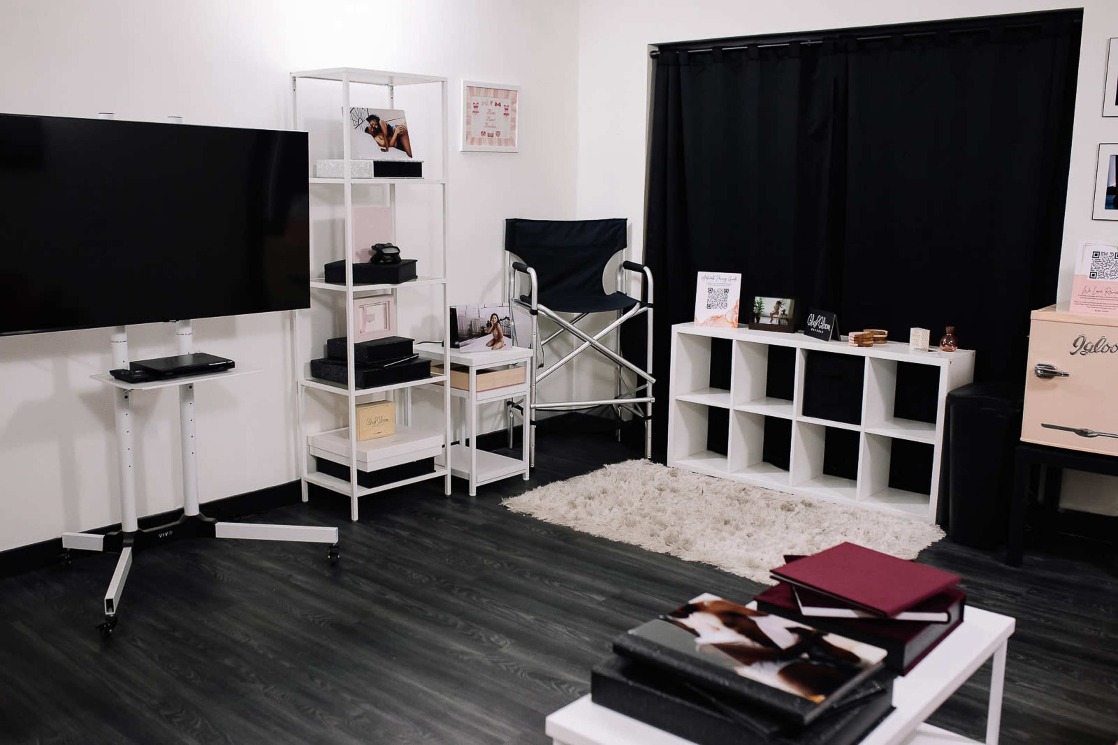 A modern, minimalist room featuring a large television on a stand, a white shelving unit with various items, a director's chair, and a stylish black curtain.