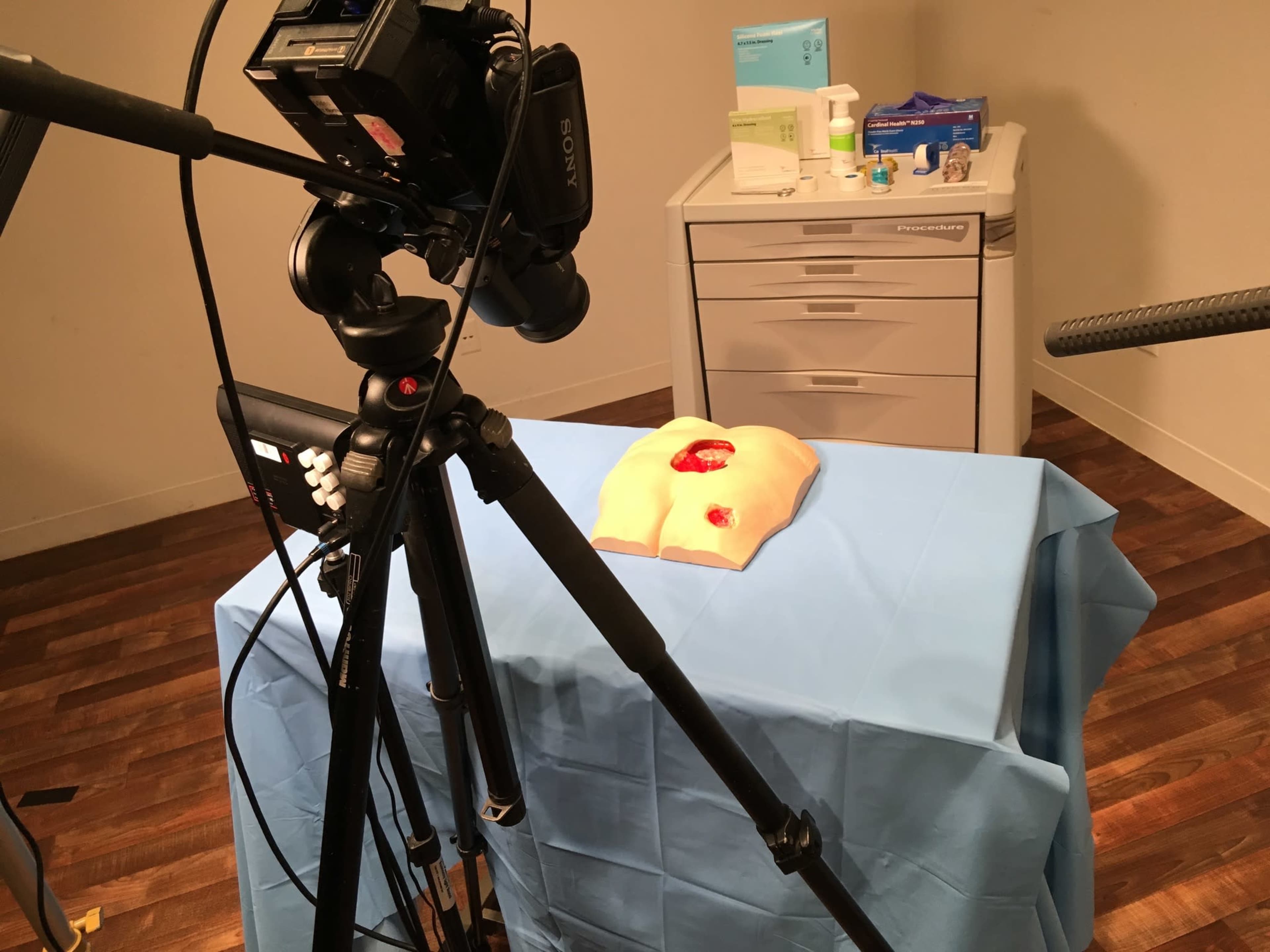 A camera is set up in front of a blue tablecloth with a medical training model and equipment on a countertop in a room.