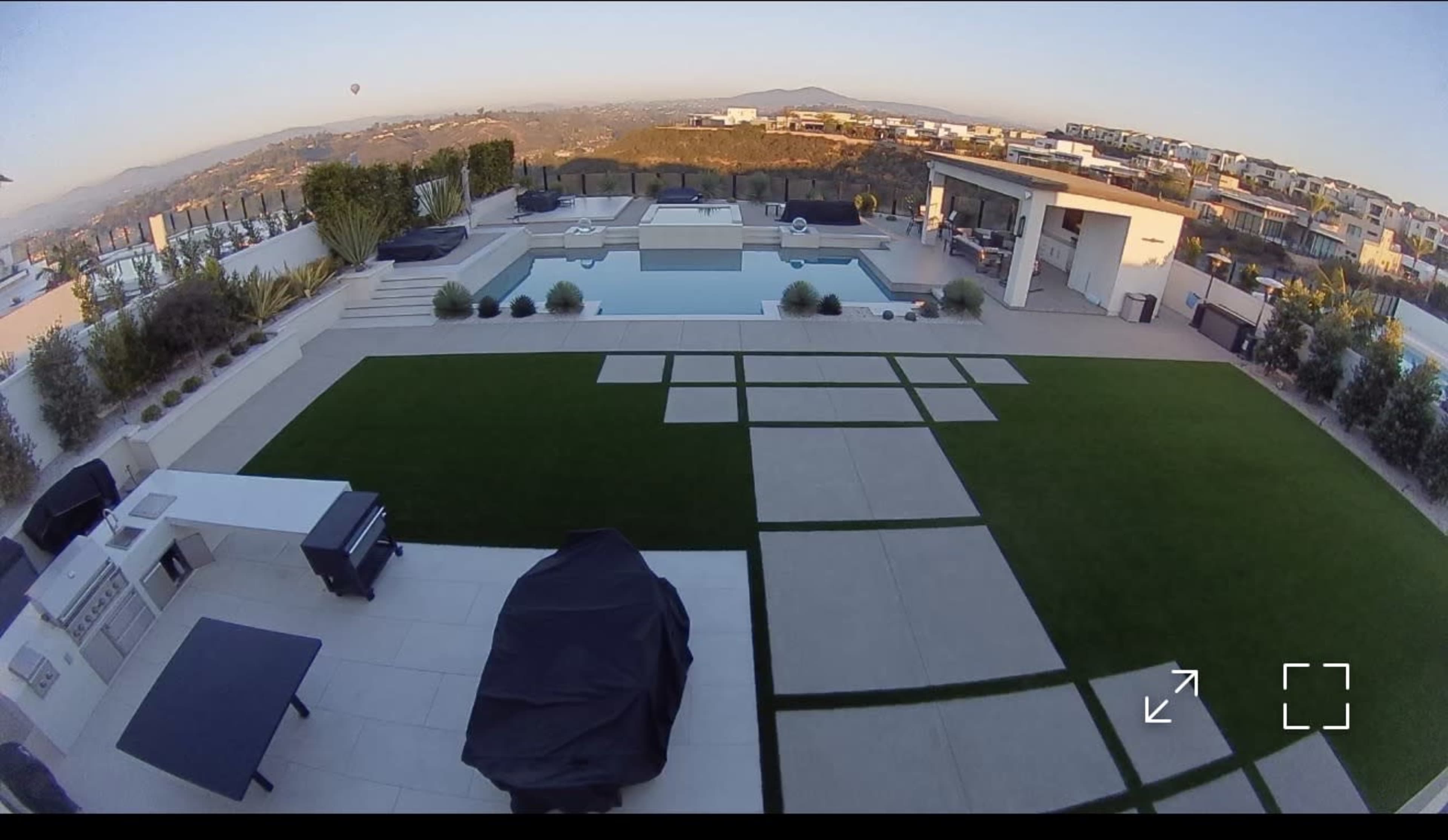 The image shows a modern outdoor patio area with a swimming pool, lounge chairs, and landscaped greenery against a backdrop of hills.