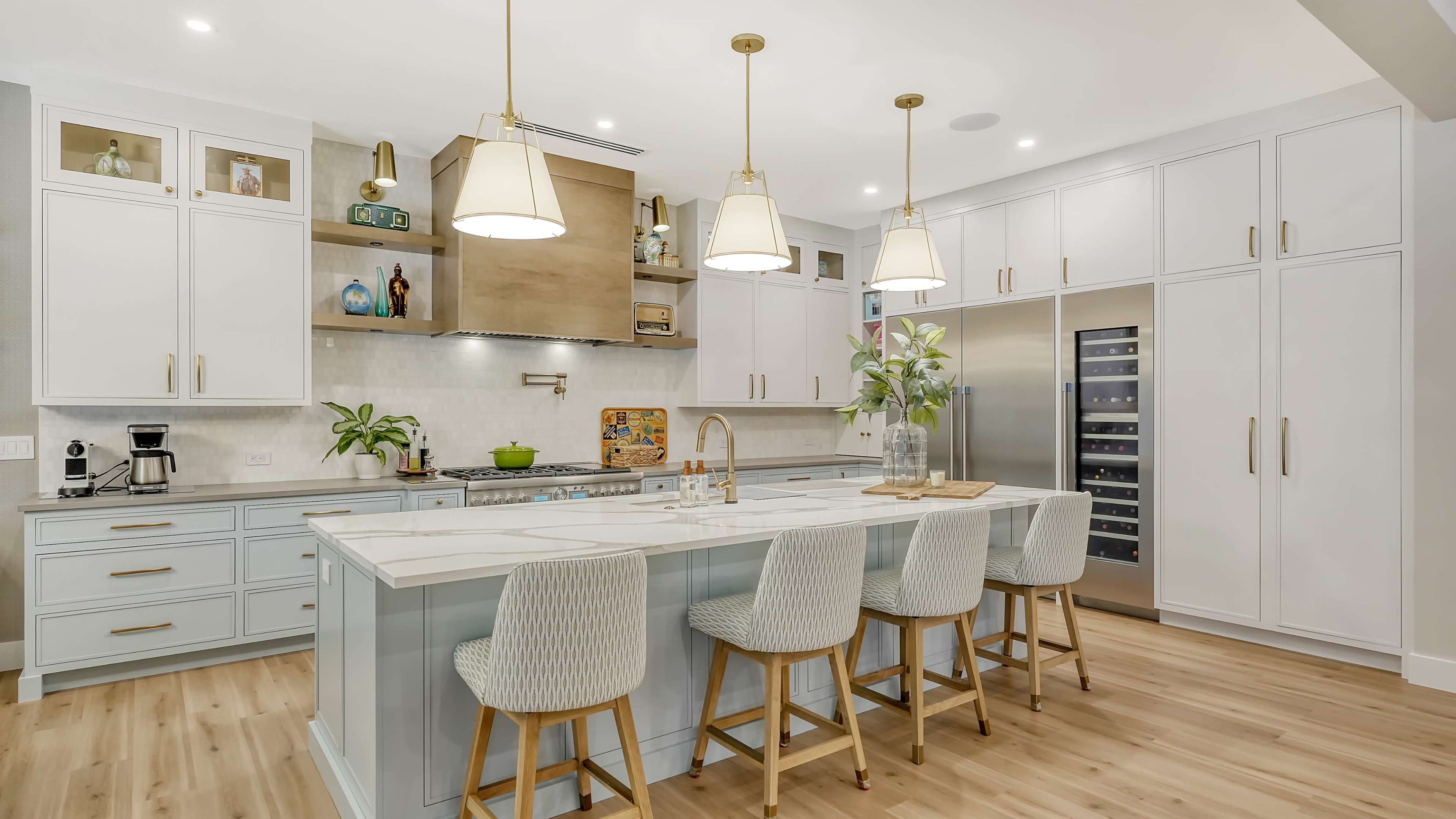 A modern kitchen features a large island with four stools, pendant lights hanging above, and sleek cabinetry alongside stainless steel appliances.