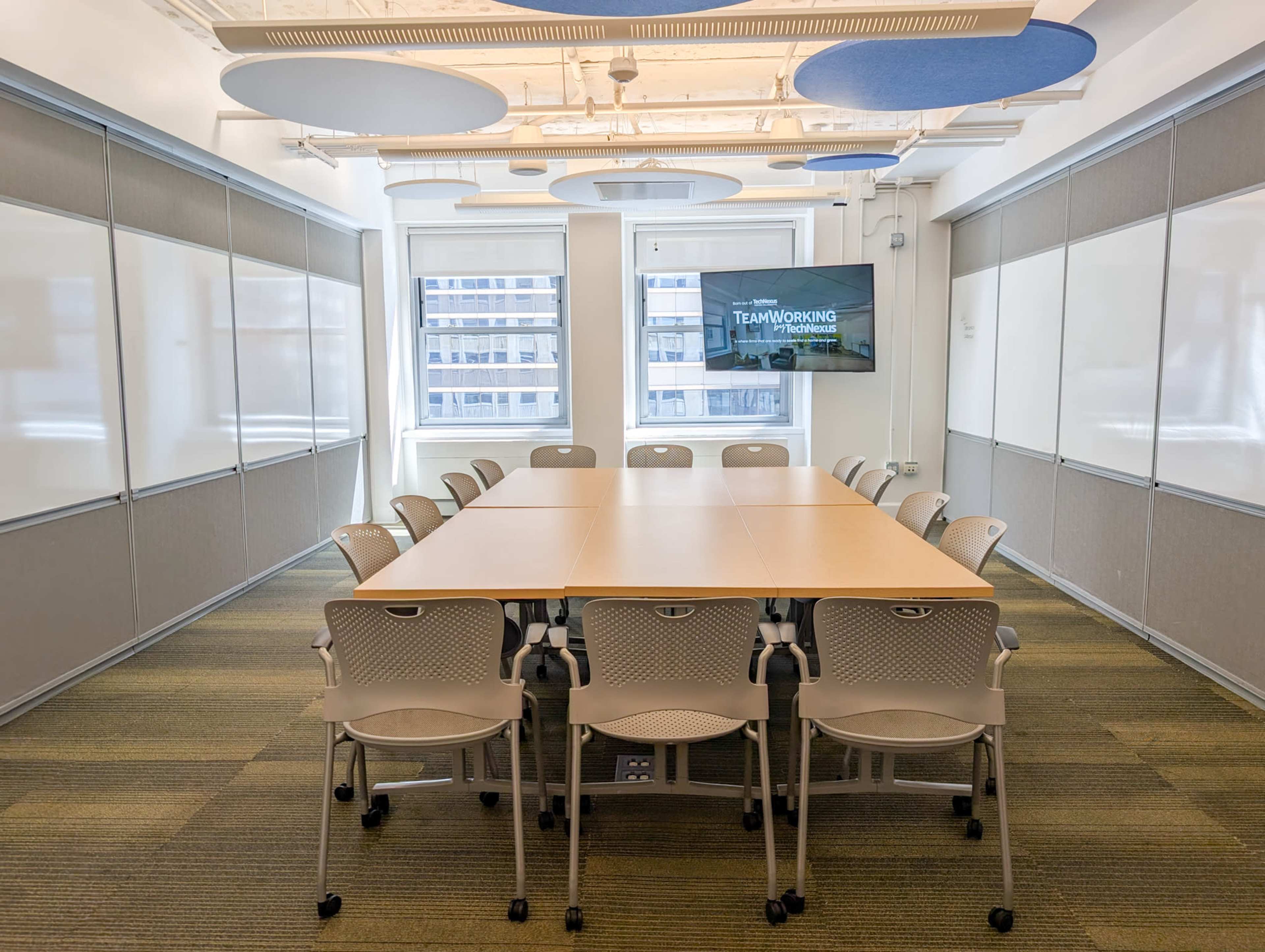 A modern conference room features a large table surrounded by rolling chairs, with whiteboards on the walls and a screen displaying the word "TEAMWORKING."