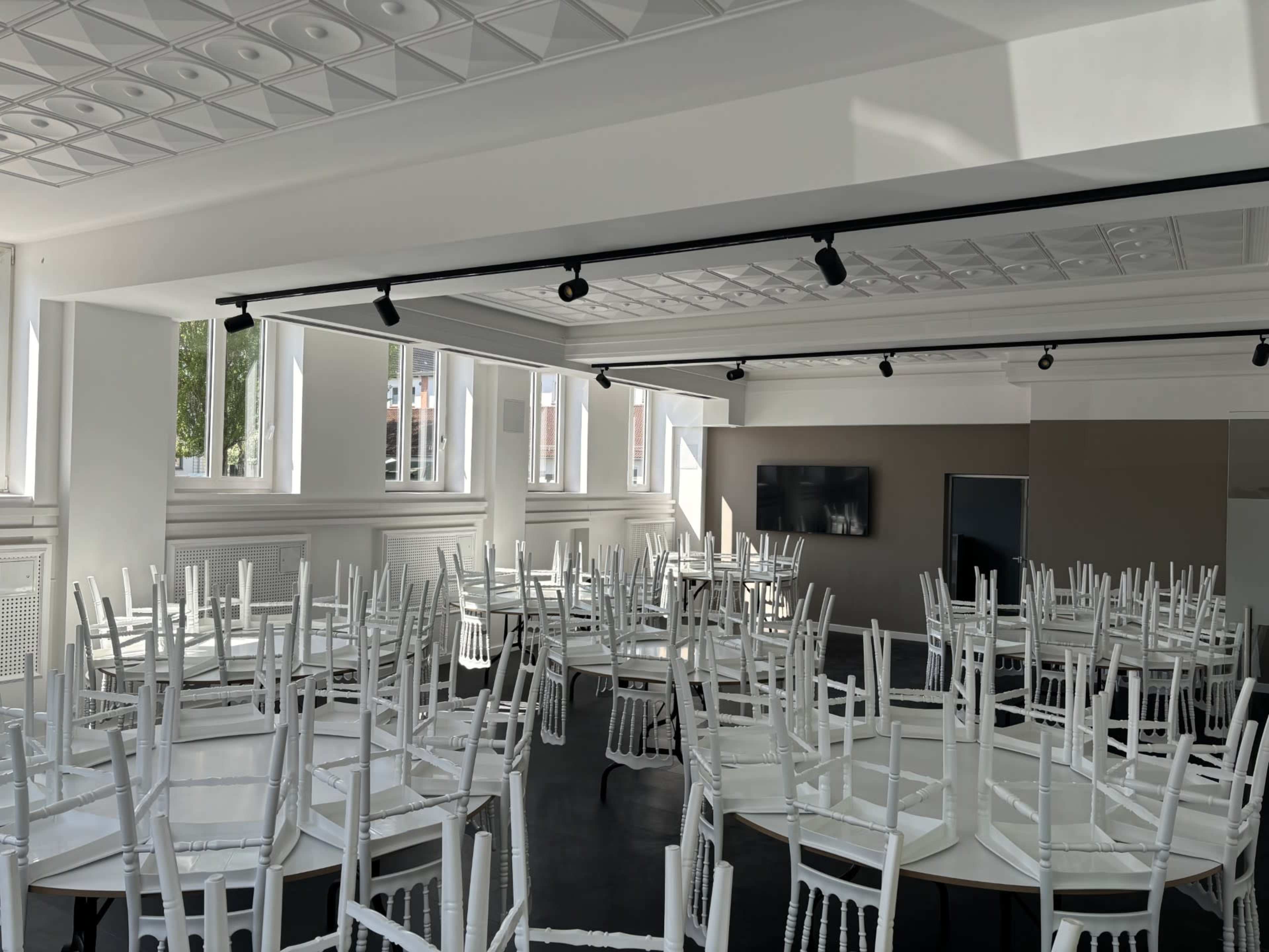 The image shows a bright, empty event space with several round tables and white chairs stacked on top.