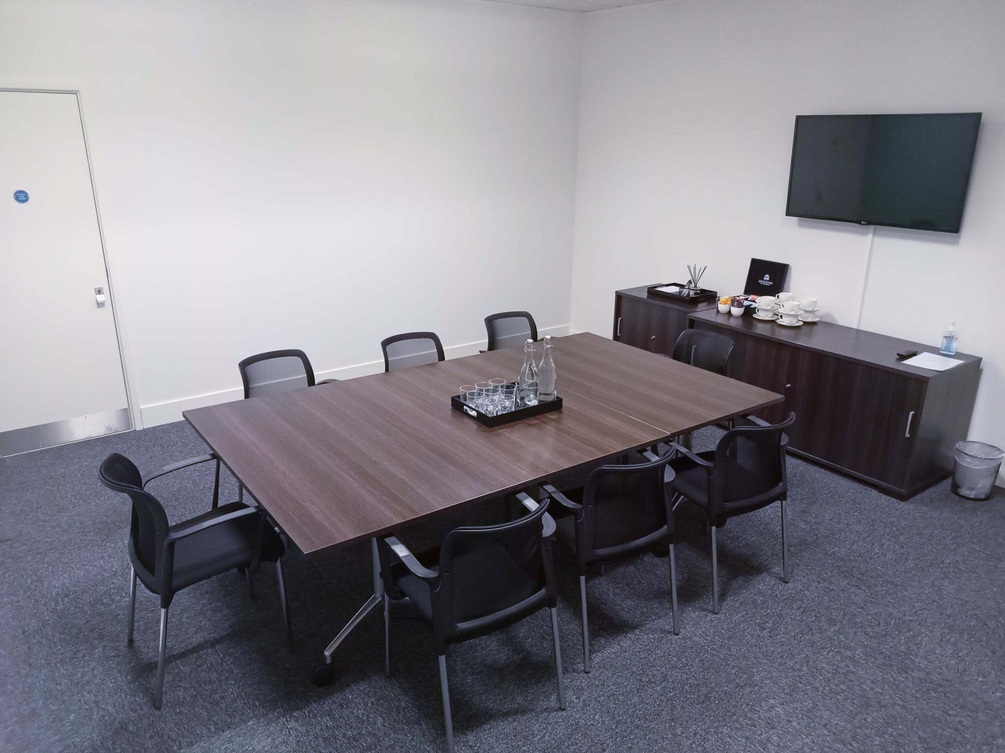 A conference room with a large table surrounded by black chairs, a television on the wall, and refreshments on a side cabinet.
